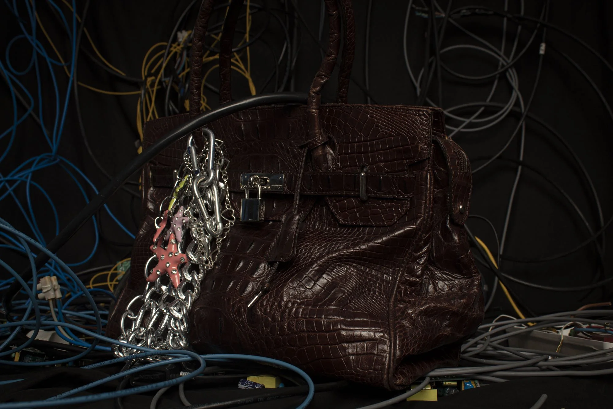 Bolso de mano de color marrón con textura de cuero de cocodrilo y llaveros colgados, rodeado de cables eléctricos enredados.