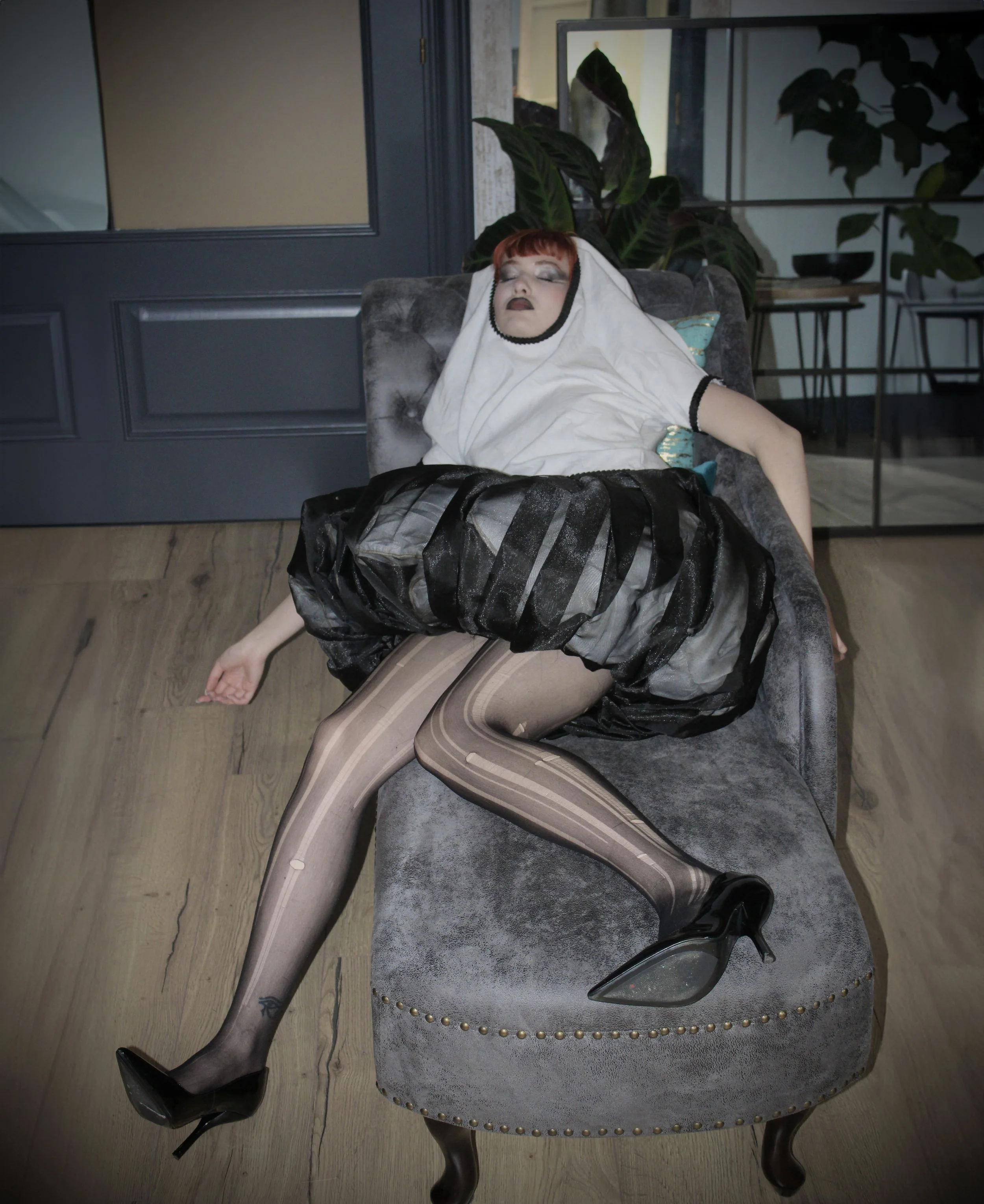 Mujer durmiendo en una silla, vestida con falda de leopardo, medias de rayas y camiseta blanca, con cabellera castaña, en un entorno interior con plantas y muebles modernos.