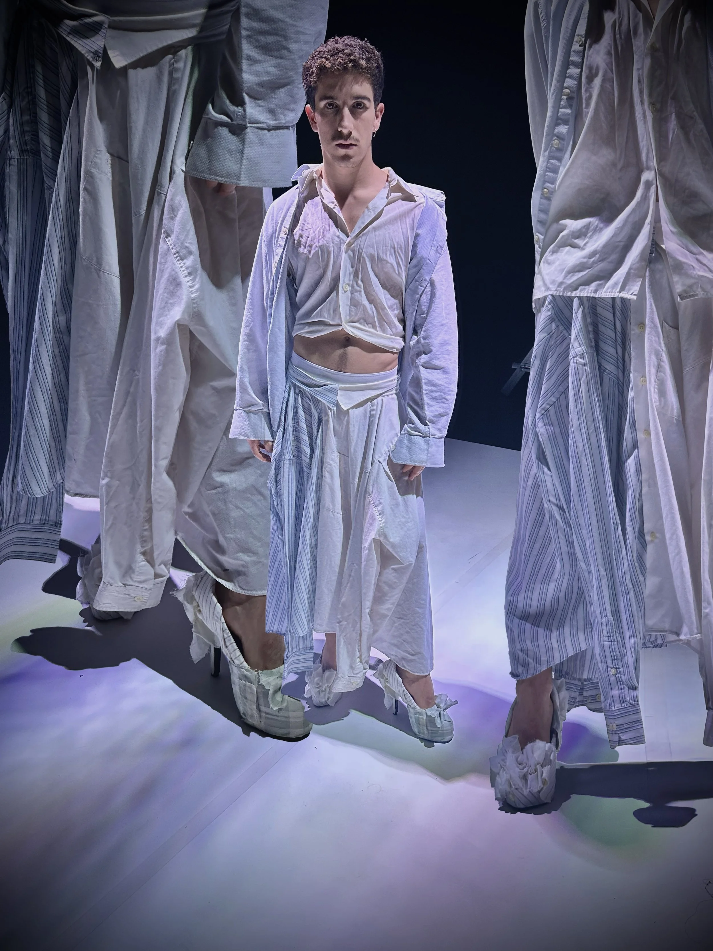Hombre con ropa blanca de pijama, rodeado de otras personas vestidas igual, en un escenario con iluminación suave.