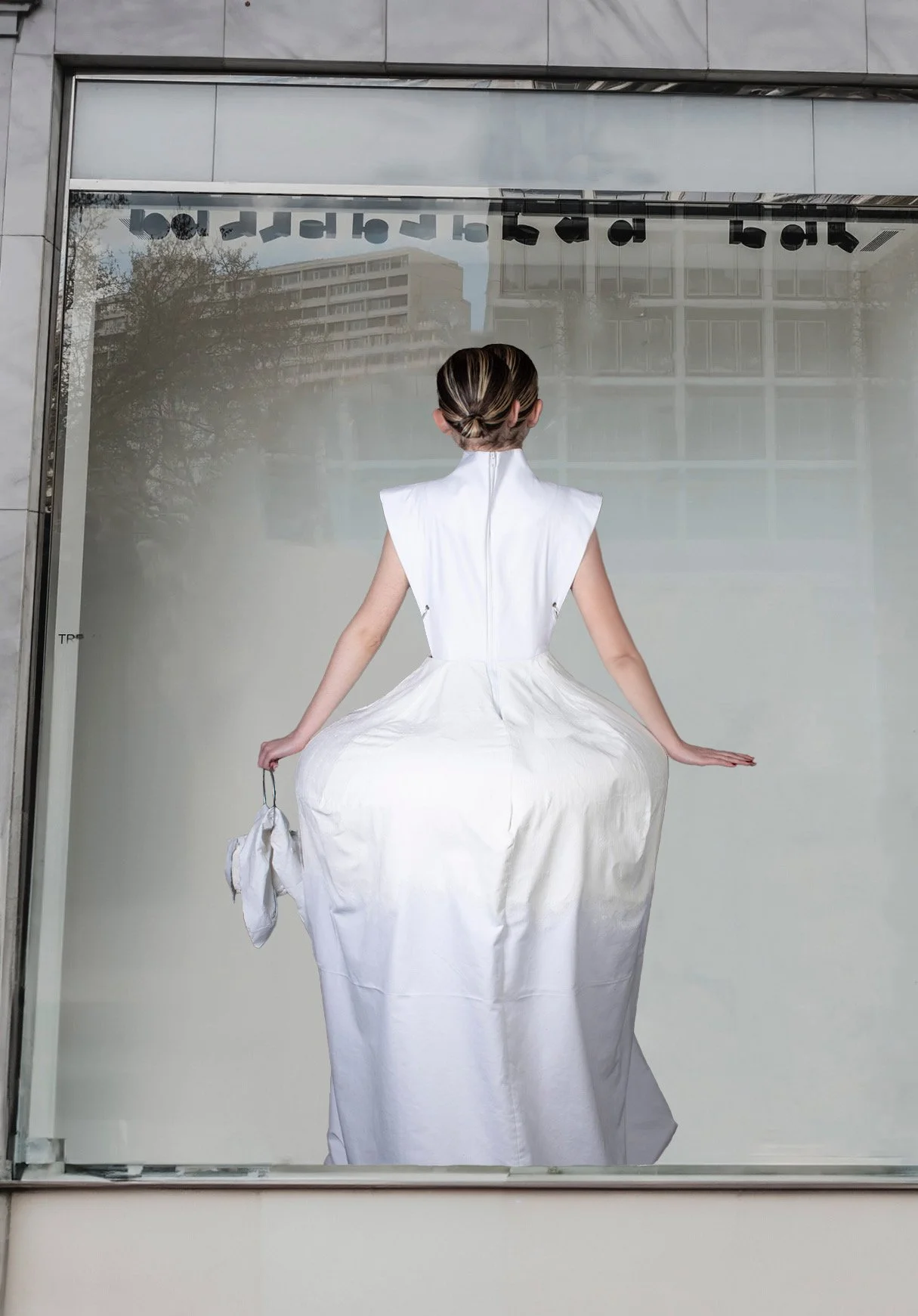 Mujer con vestido largo y peinado en forma de moño, vista de espaldas, en una tienda con cristal reflectante y edificios en el fondo.