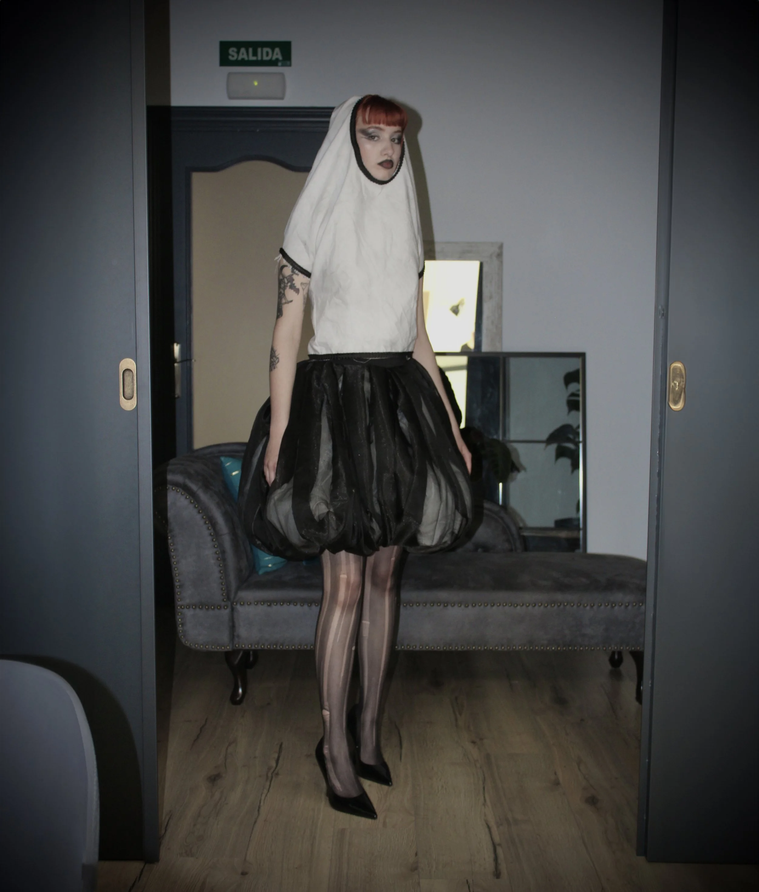 Mujer con rostro maquillado y cabello rojo, vestida con una prenda que imita un saco de papas y falda larga de tul negro, con pantimedias de rayas y tacones negros, de pie frente a un espejo en una habitación con paredes grises y una puerta con señal