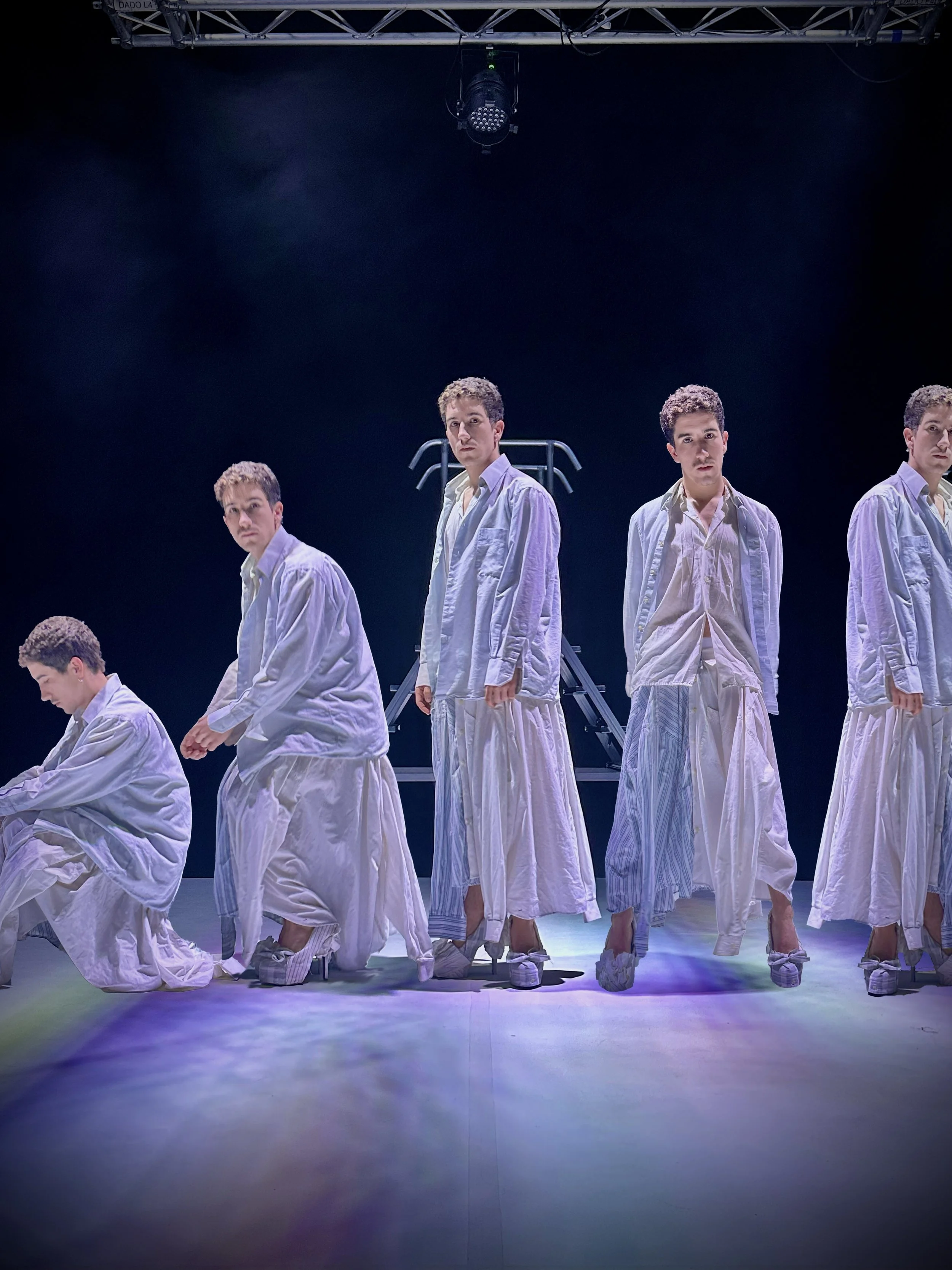 Un grupo de siete personas con ropa blanca, algunas están de pie y otras están en cuclillas sobre el escenario, en una puesta en escena teatral o de danza, en un fondo oscuro con iluminación que destaca a las personas.
