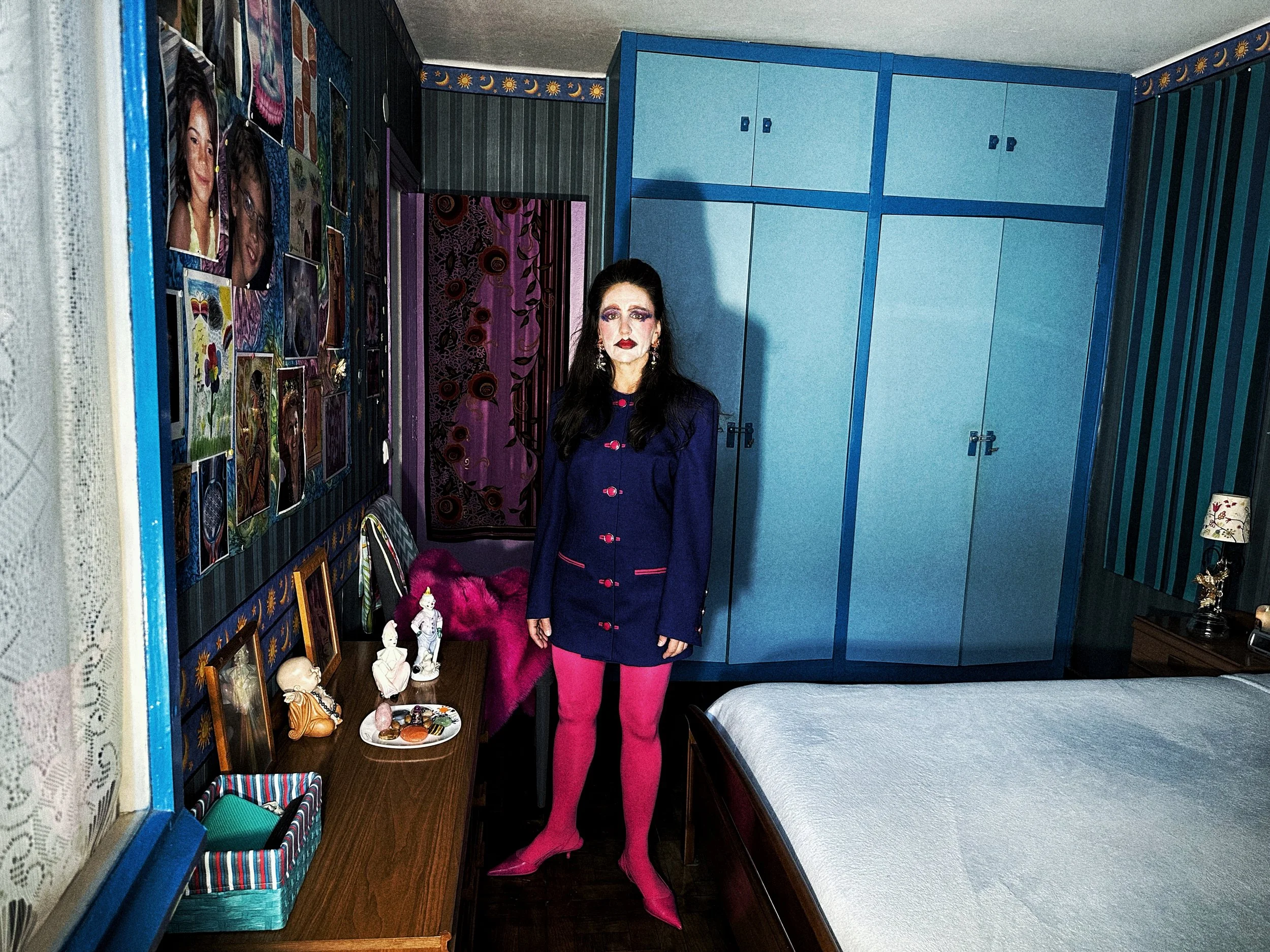 Una mujer con maquillaje llamativo, ropa azul y pantalones rosa, de pie en un dormitorio con paredes decoradas con fotos y cuadros. Frente a un armario azul y junto a una cama, en una habitación decorada con diversos objetos y arte.