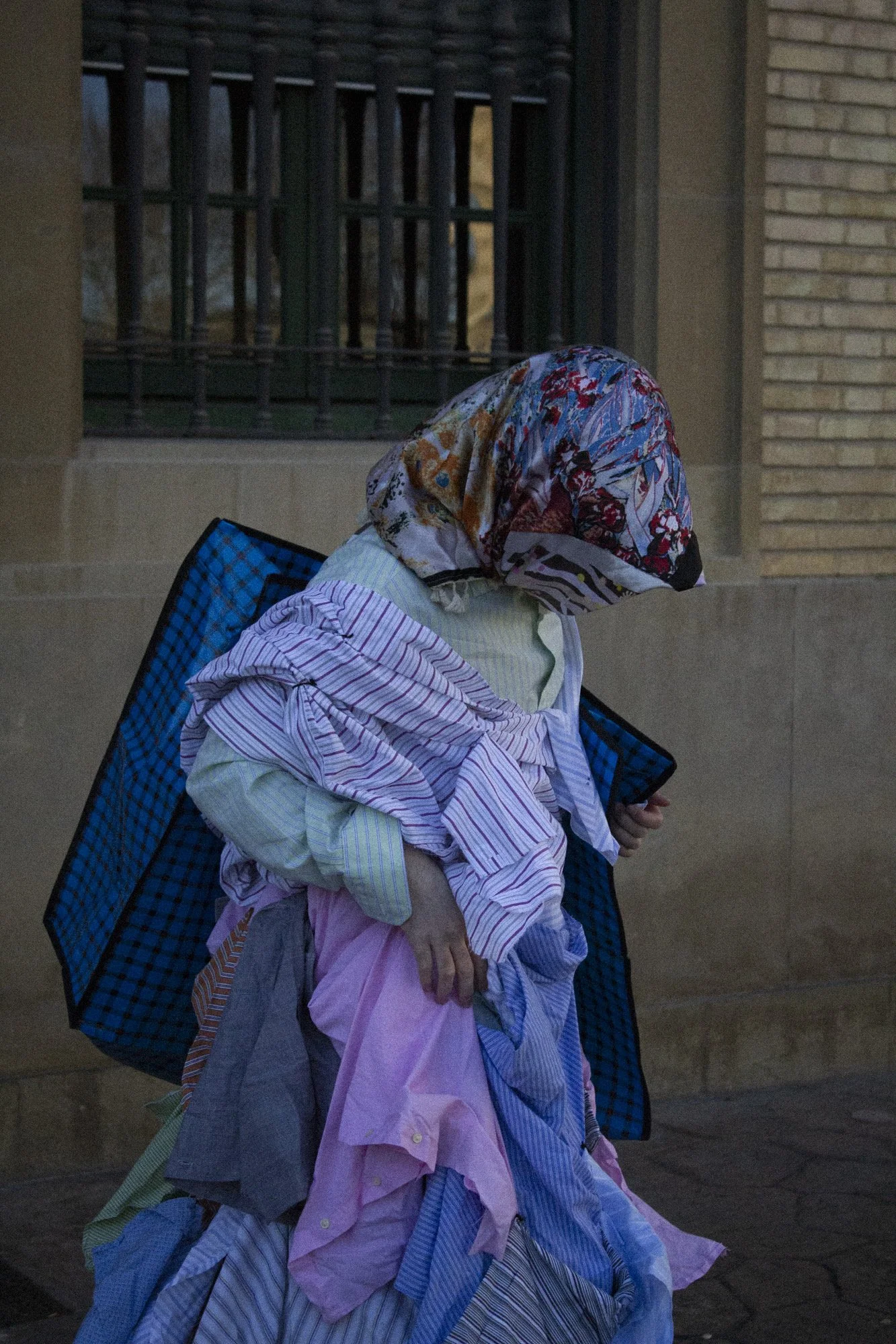 Persona con ropa y pañuelo en la cabeza, sosteniendo una bolsa, caminando cerca de un edificio con muro de ladrillos y una reja
