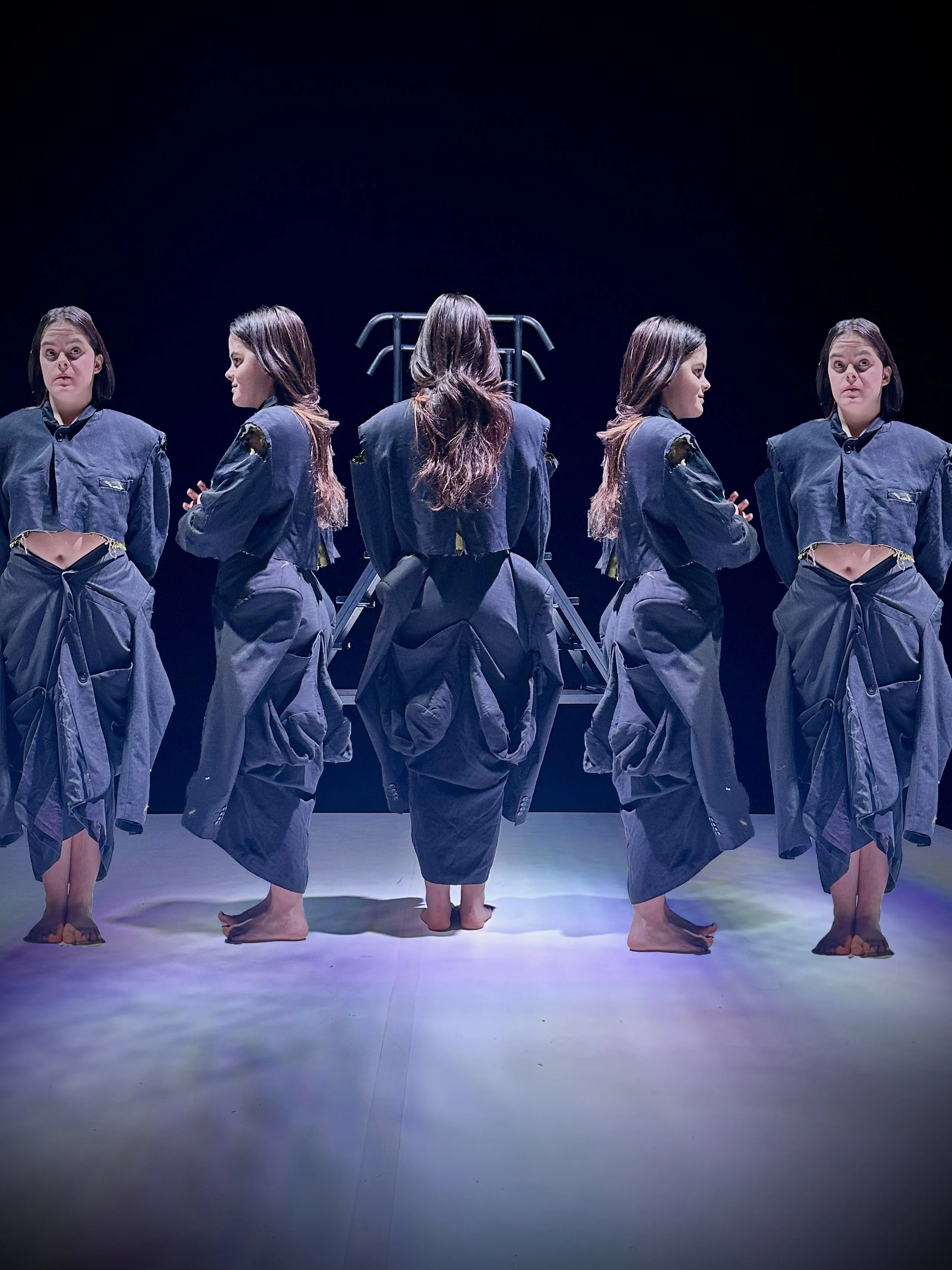 Seis mujeres en ropa azul, de espaldas o de perfil, que parecen estar en un escenario con fondo oscuro. Algunas están mirando hacia adelante, otras hacia los lados, y una más tiene los ojos cerrados. Están de pie, sin zapatos, en diferentes posicione