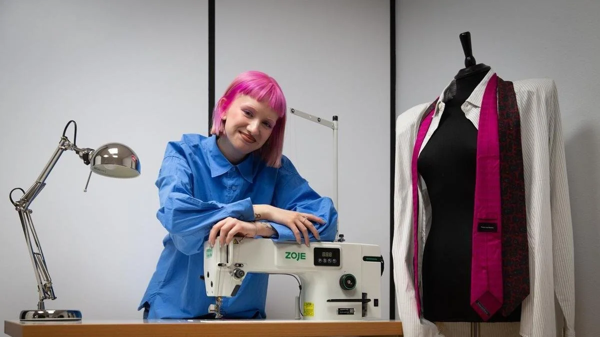 Joven con cabello rosa usando camisa azul en un taller de costura, con máquina de coser y vestido en maniquí