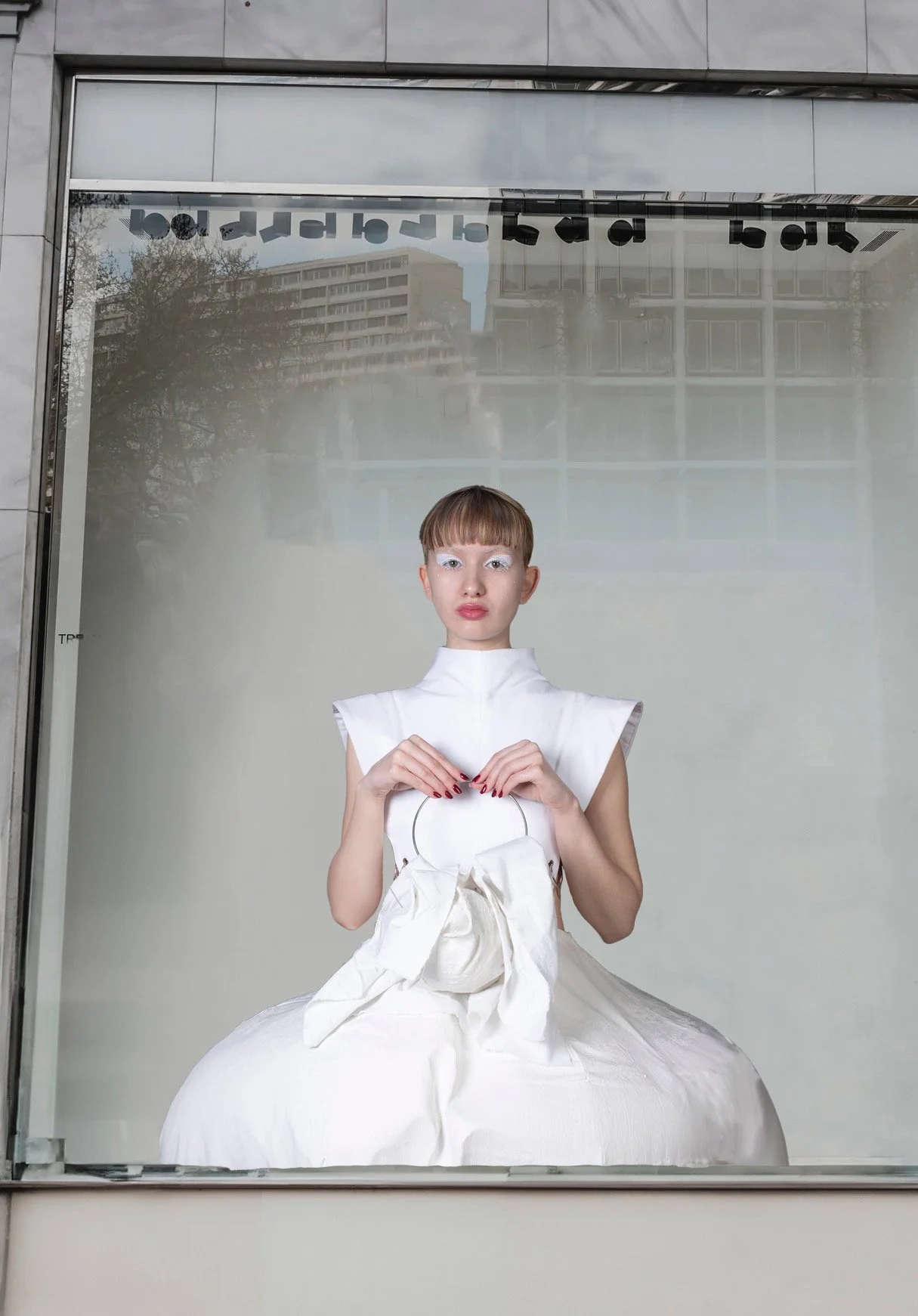 Mujer con maquillaje blanco en los ojos y vestido blanco, sosteniendo una bolsa blanca, en una ventana de la ciudad.