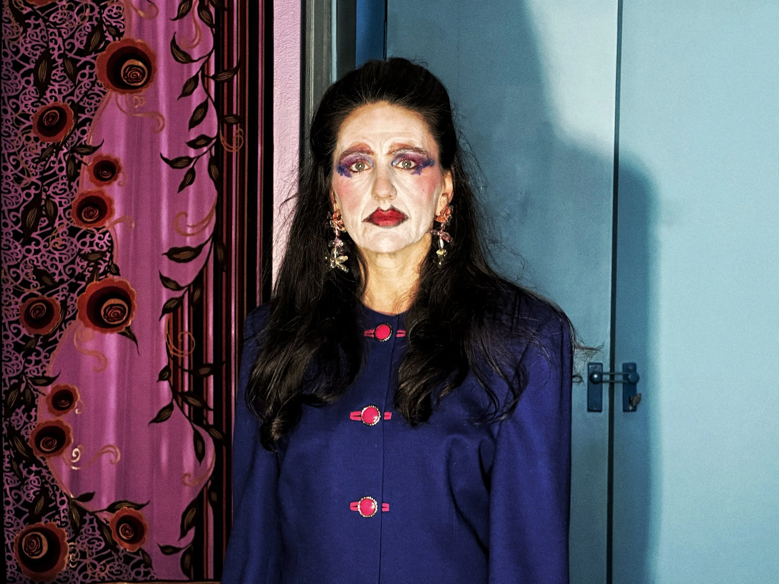 Una mujer con maquillaje artístico, orejas prominentes, cabello oscuro y largo, viste un vestido azul con detalles rosados y aretes grandes, posando frente a una pared con cortina rosa con flores y un fondo azul.