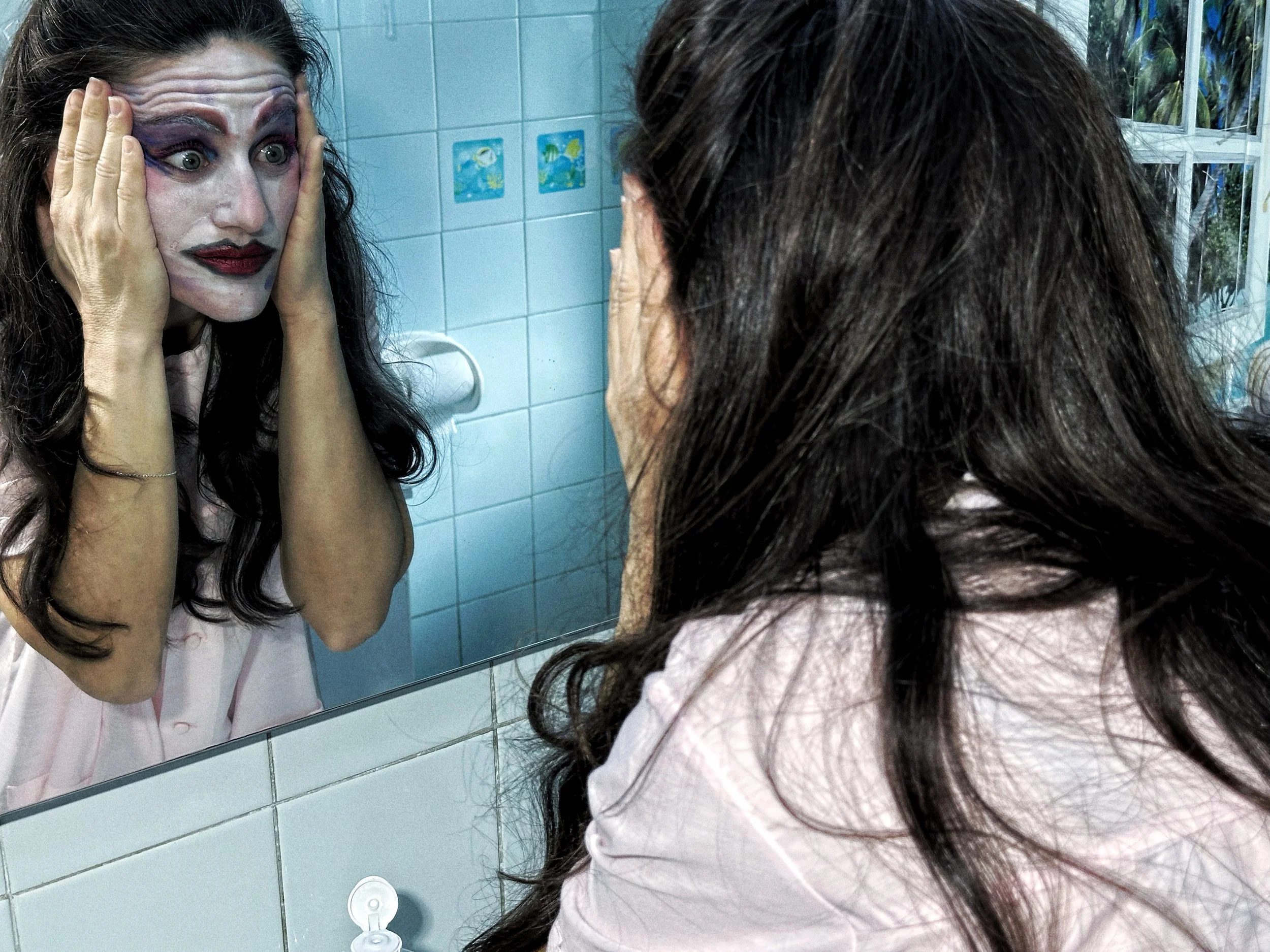 Mujer con maquillaje de payaso en el rostro, viendo su reflejo en el espejo del baño, mientras sostiene sus mejillas con ambas manos.