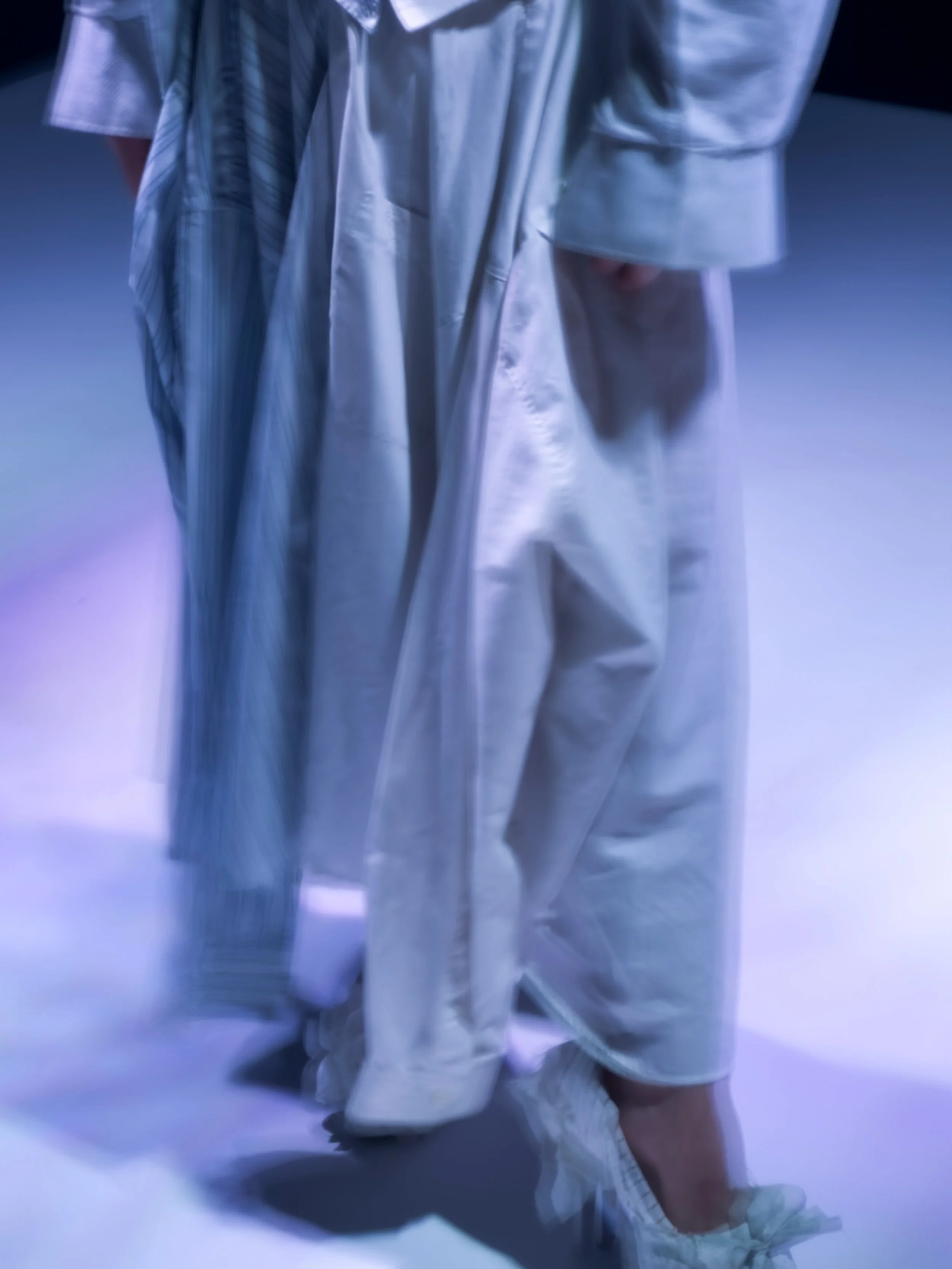 Persona vestida con un traje plateado y zapatos deportivos blancos, al parecer en una sesión de fotos o en un escenario con fondo de tonos oscuros y luz tenue.