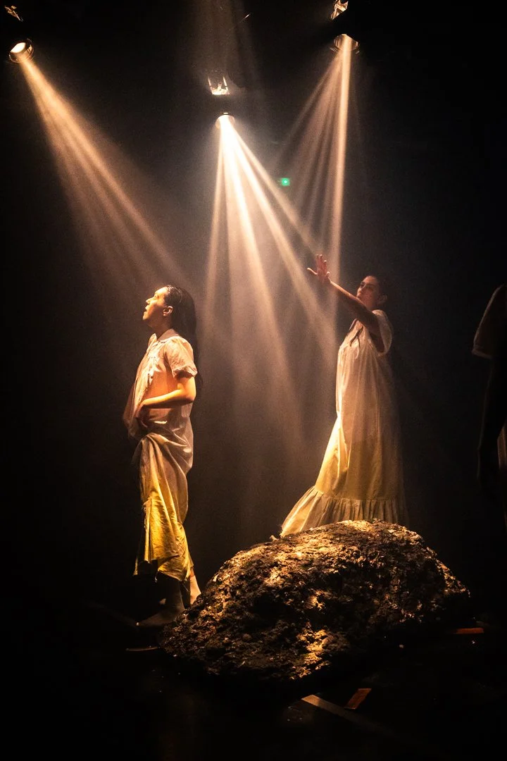 Dos personas en un escenario con luces de escenario, una de ellas de espaldas y la otra con brazo extendido, ambas con vestidos largos y sentadas sobre una roca.