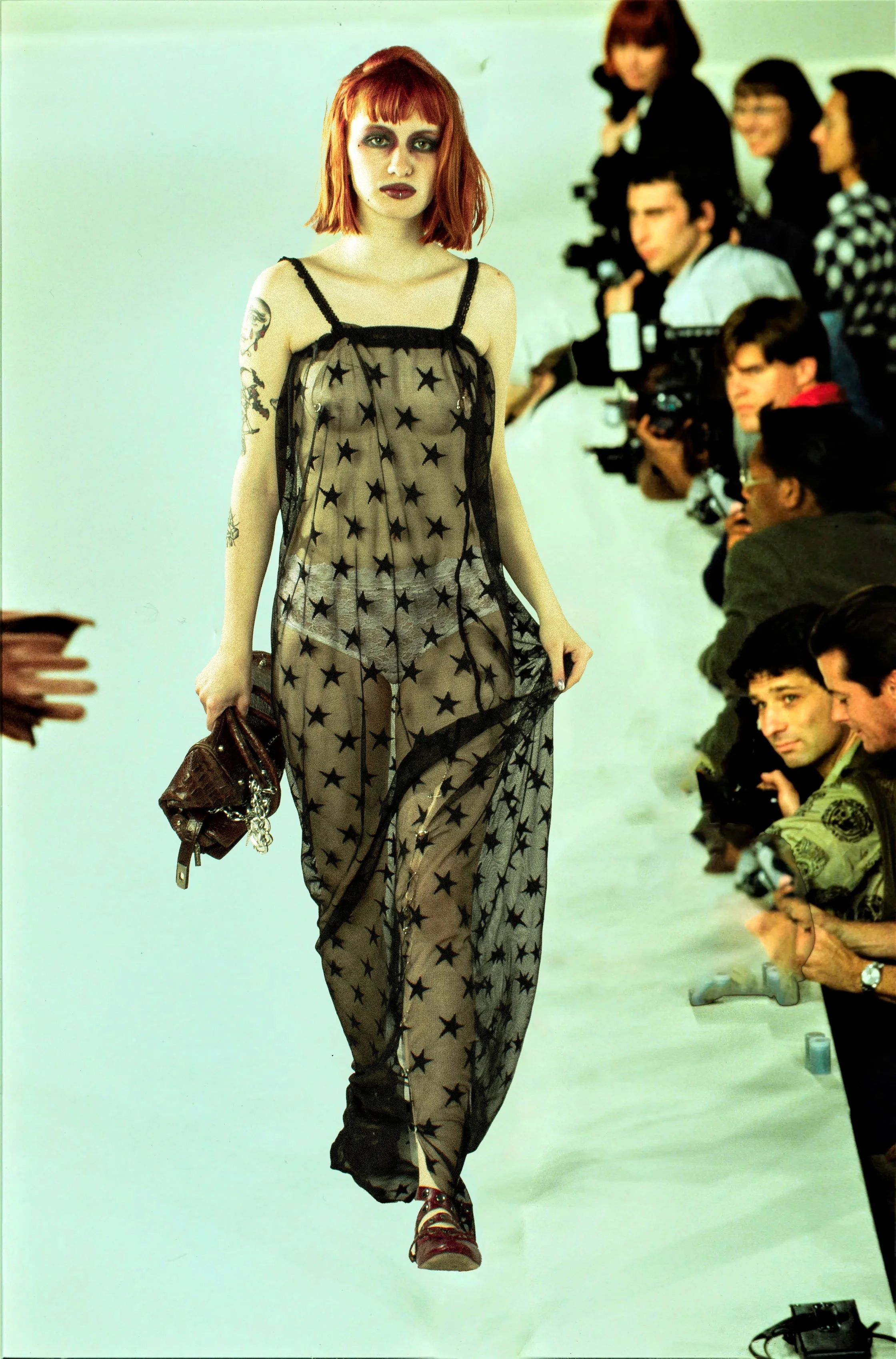 Modelo caminando en pasarela con fondo de personas y fotógrafos, vestido de malla con estrellas y pantalones transparentes, peinado de cabello rojo corto y maquillaje oscuro.