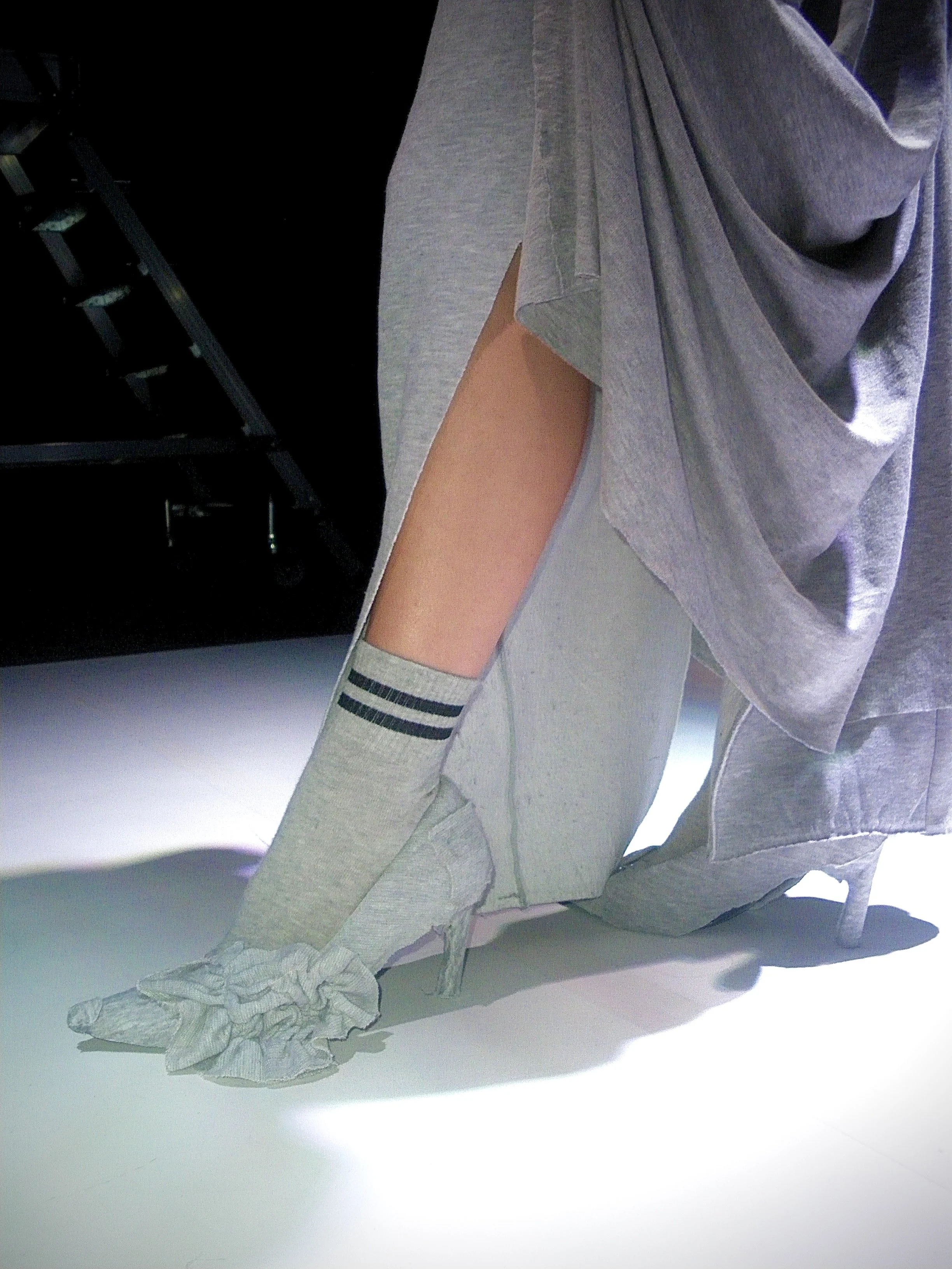 Persona vestida con ropa cómoda y calcetas, sentada en un piso blanco, con una estructura metálica y objetos en el fondo.