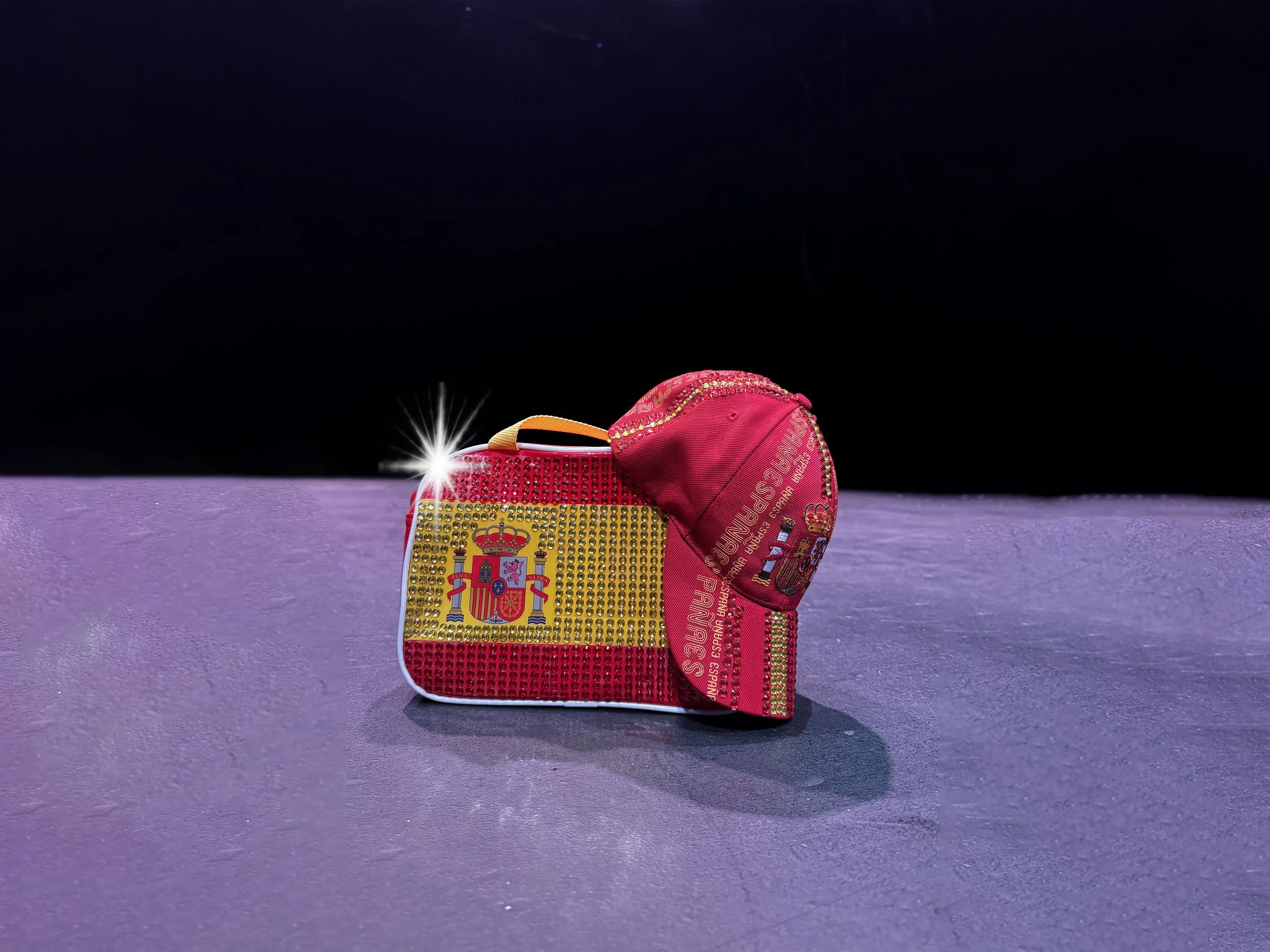 Un pequeño bolso en forma de maleta decorado con cristales dorados y simbolos de la selección española, acompañado de una gorra rosa con detalles en dorado y el escudo de España.