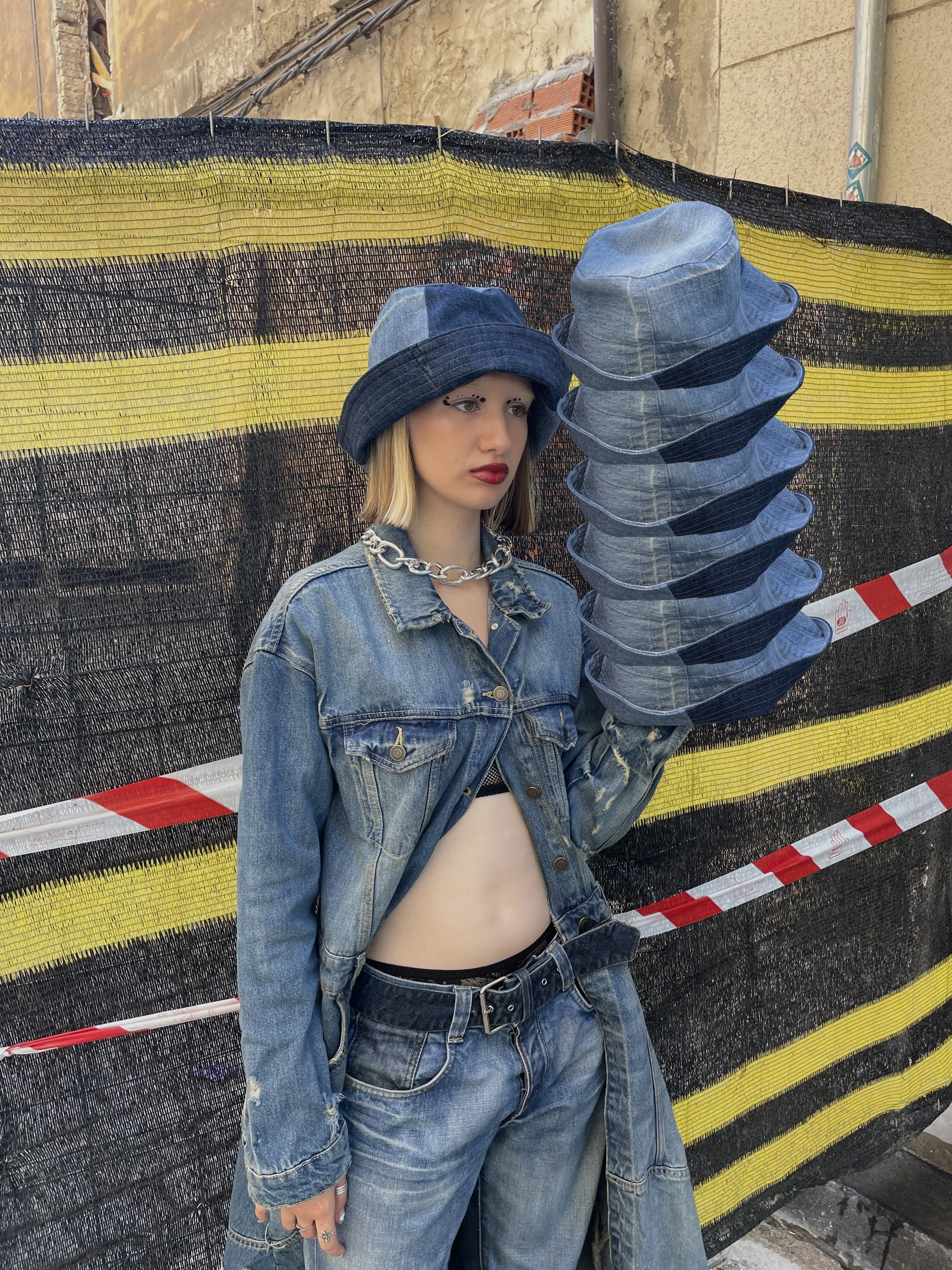 Adolescente con gorro de mezclilla, chaqueta y pantalones de mezclilla, de cabello rubio y corto, posando junto a una estructura de lona negra y amarilla con una cinta de seguridad roja y blanca.