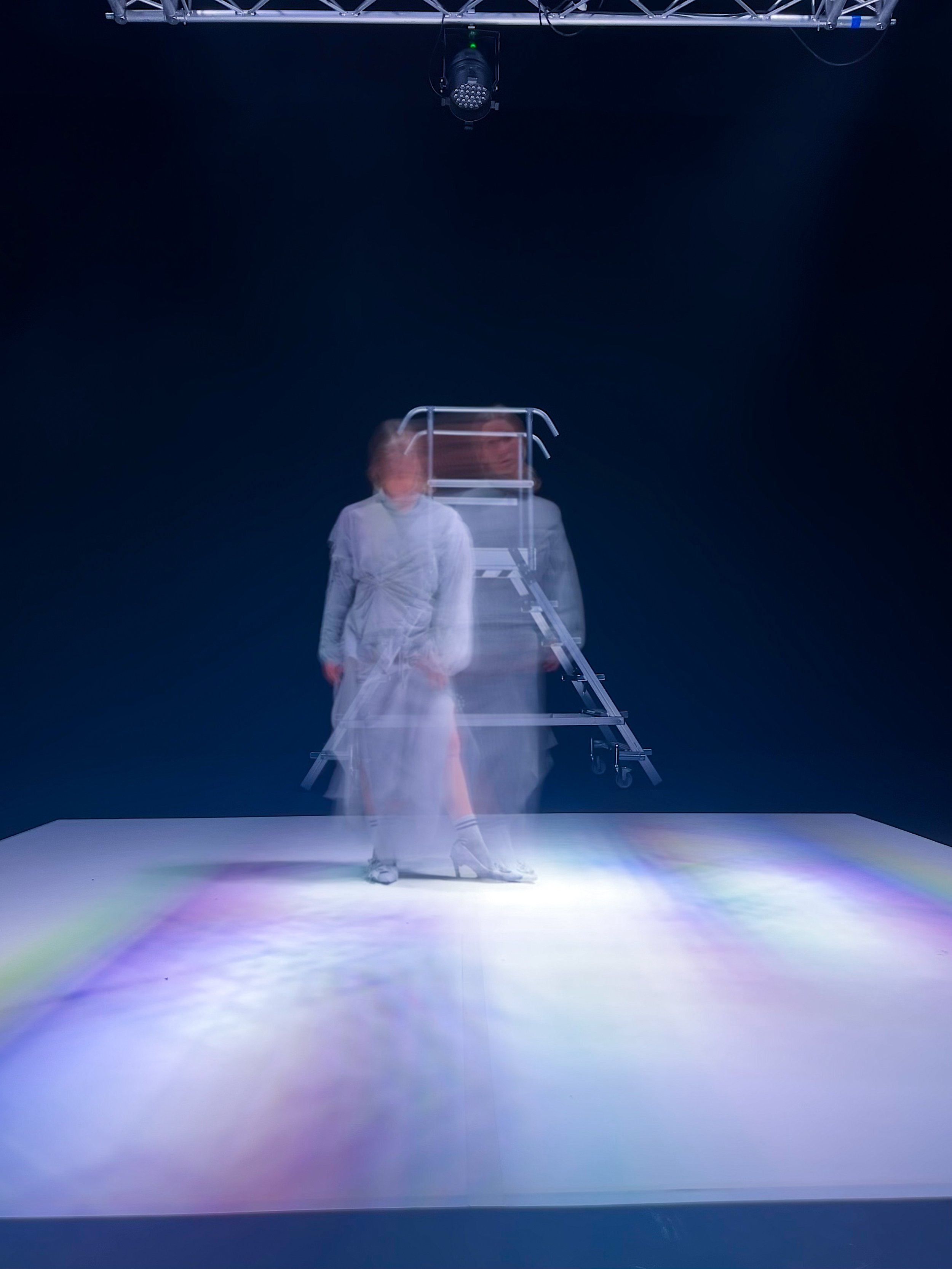 Figura de una persona con prendas blancas, en un escenario oscuro, con un efecto de doble exposición que crea una ilusión de movimiento, en un fondo oscuro con iluminación en el suelo que produce un arco iris.