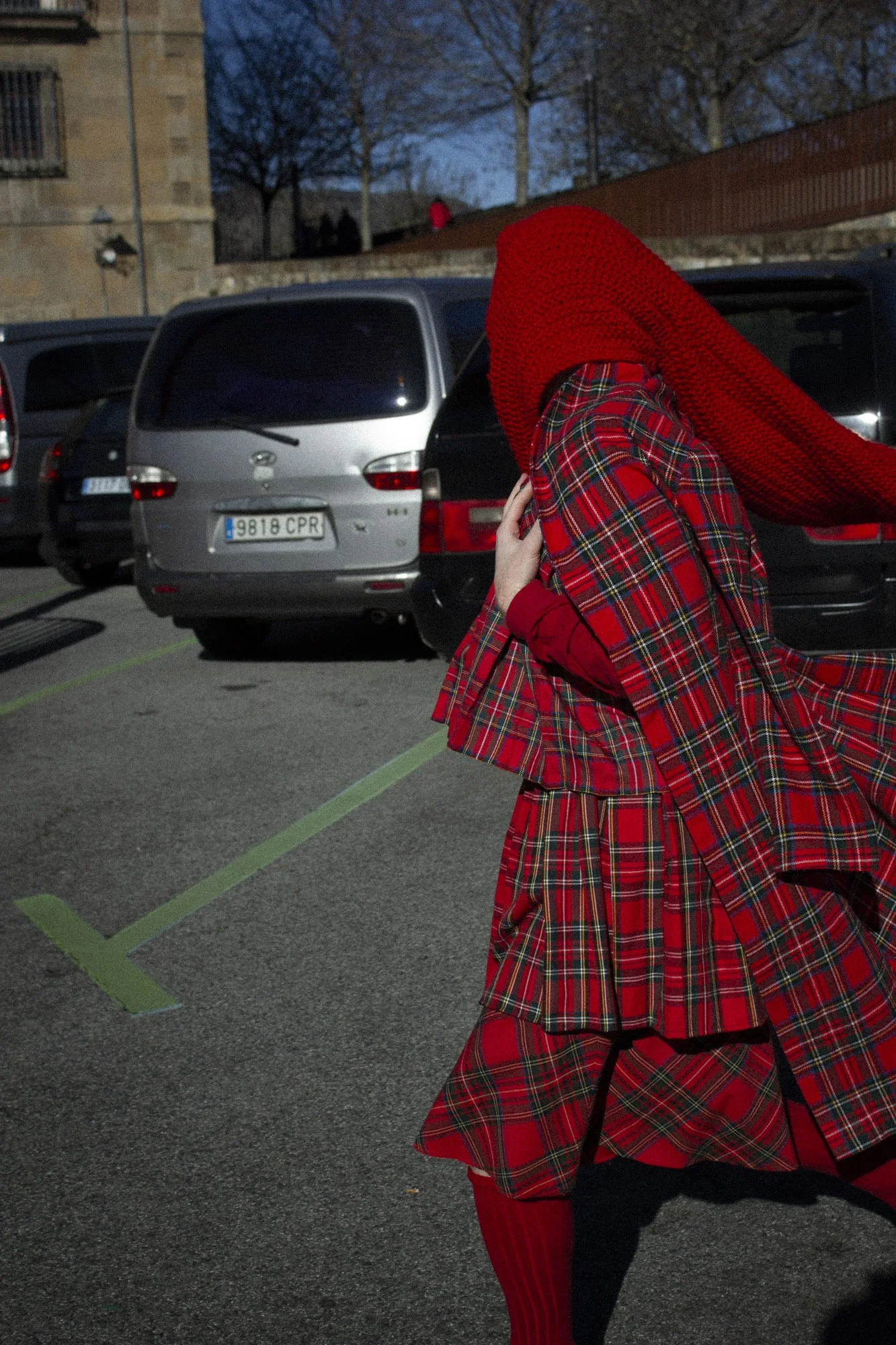Persona disfrazada con ropa de tartán rojo, usando un sombrero de punto del mismo color, en un estacionamiento por la tarde, con autos estacionados y árboles sin hojas en el fondo.