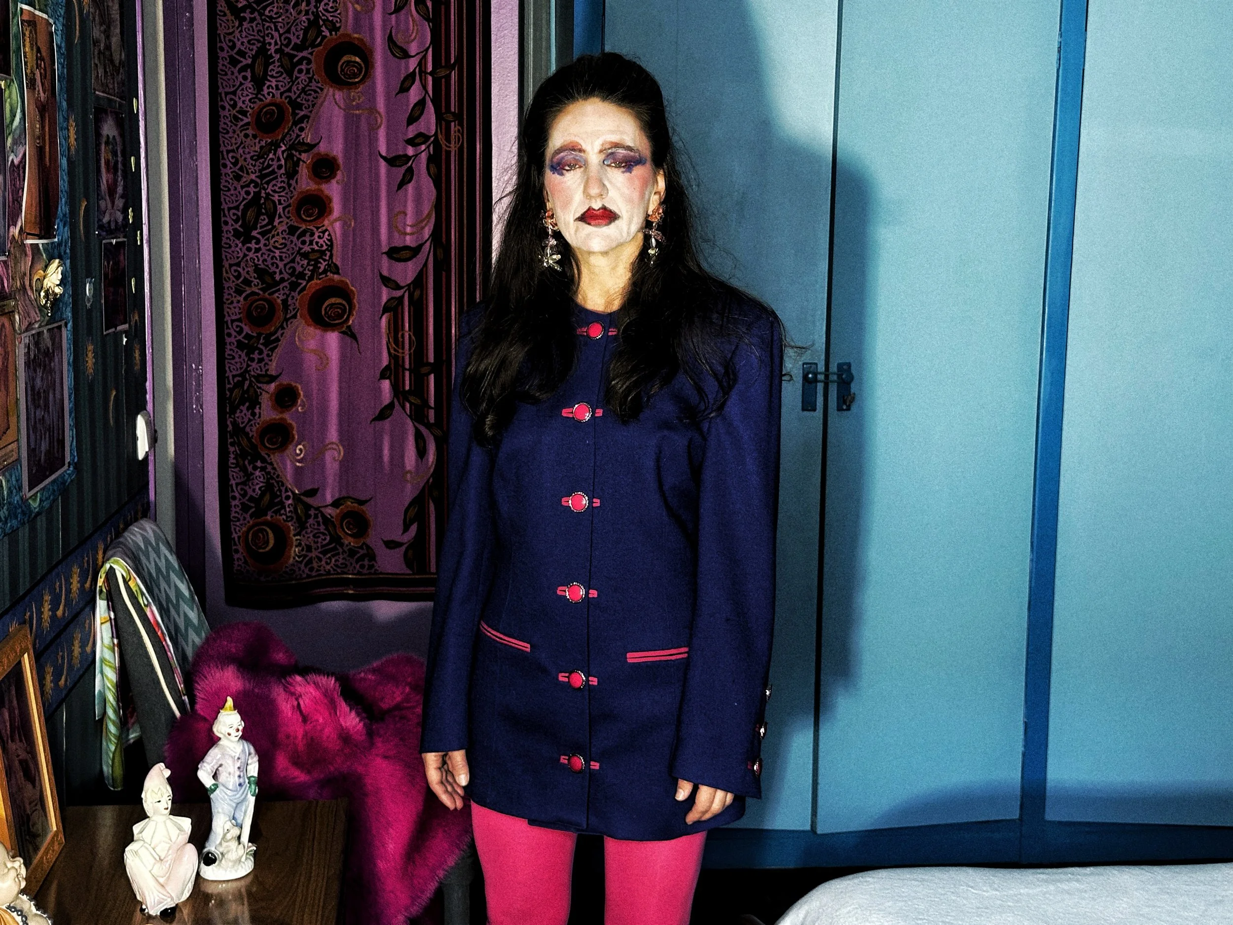 Persona vestida con un abrigo azul oscuro decorado con botones rosados, con maquillaje llamativo y pendientes grandes, posa en un cuarto decorado con telones de fondo coloridos y figuras artísticas.