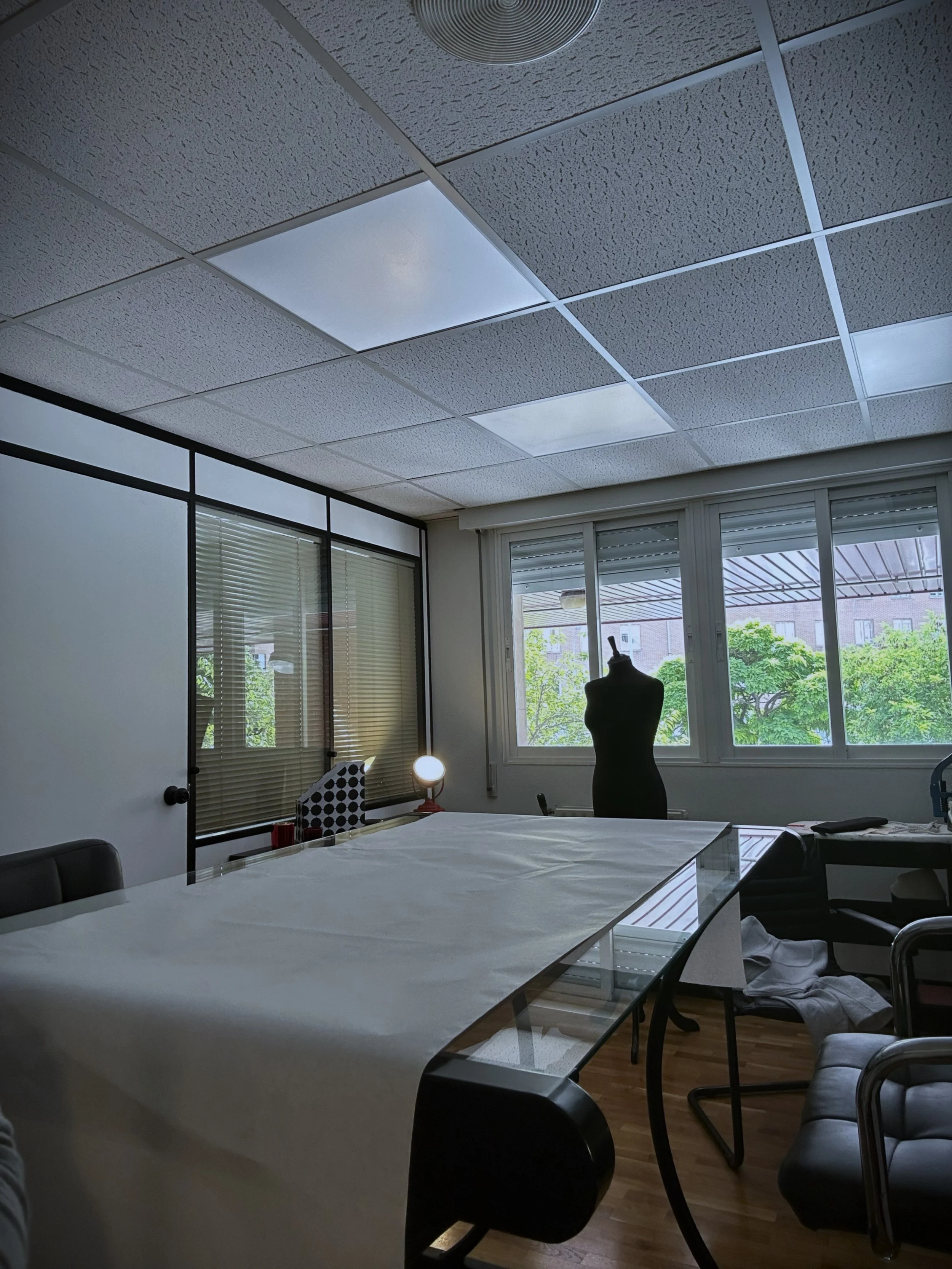 Sala de reuniones con mesa grande cubierta con tela blanca, sillas alrededor, maniquí de vestido en una esquina, ventanas con vista a árboles y edificio exterior, lámparas en el techo, persianas abiertas.