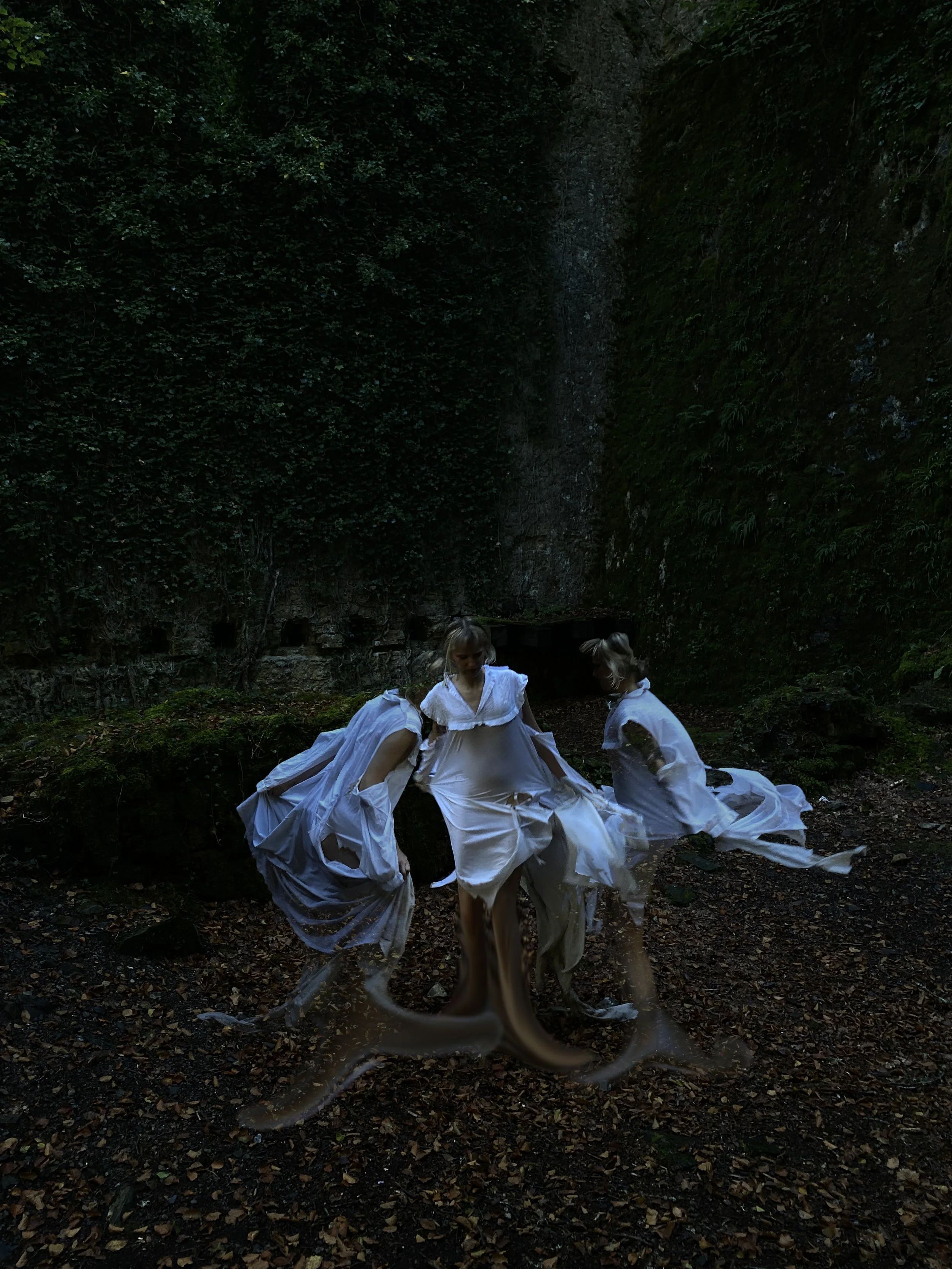 Tres niñas con vestidos blancos en un bosque, rodeadas de árboles y hojas en el suelo.