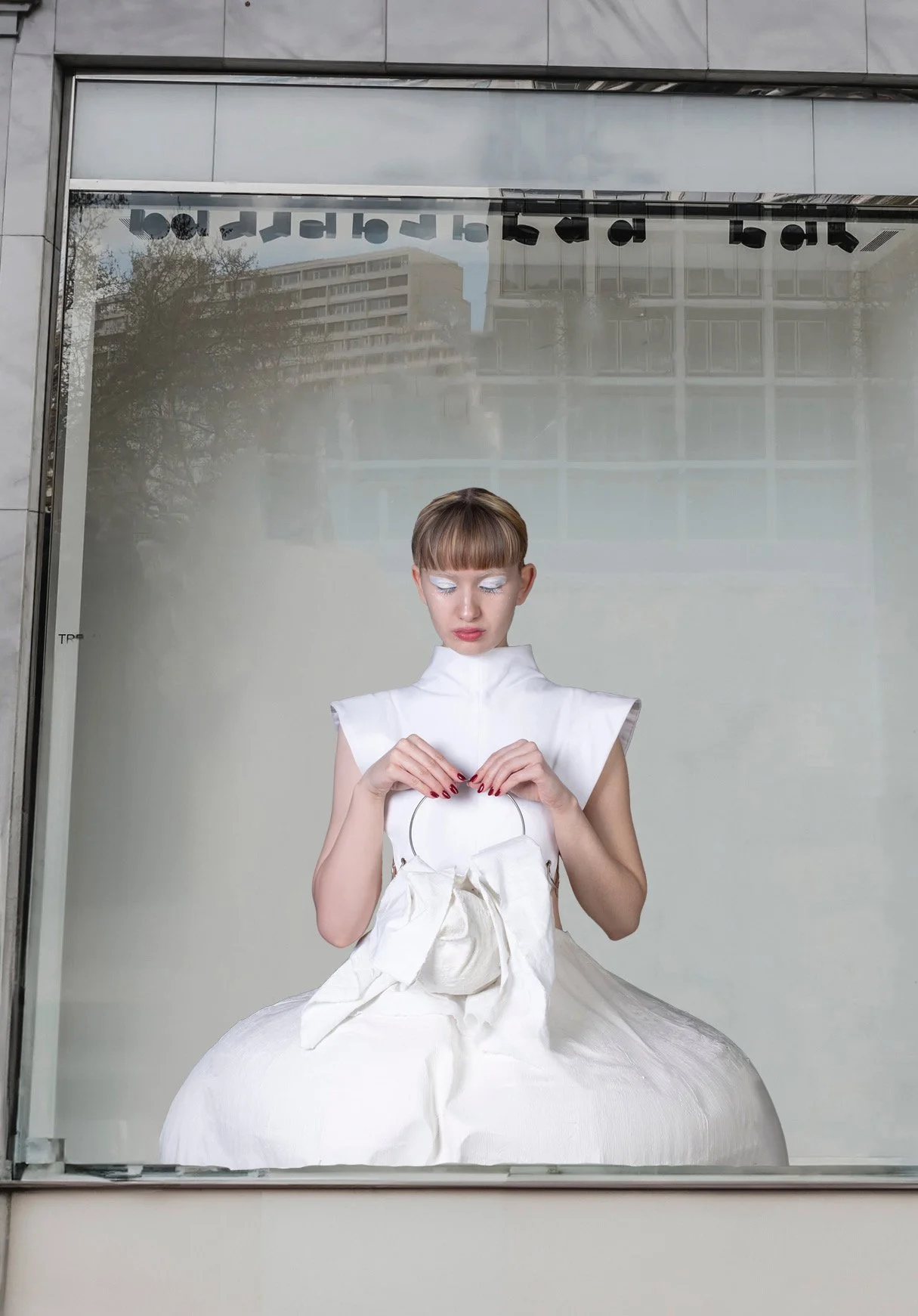Mujer con vestido blanco y maquillaje futurista, sosteniendo un bolso en forma de círculo, frente a una ventana de vidrio con reflejo de edificios y árboles.