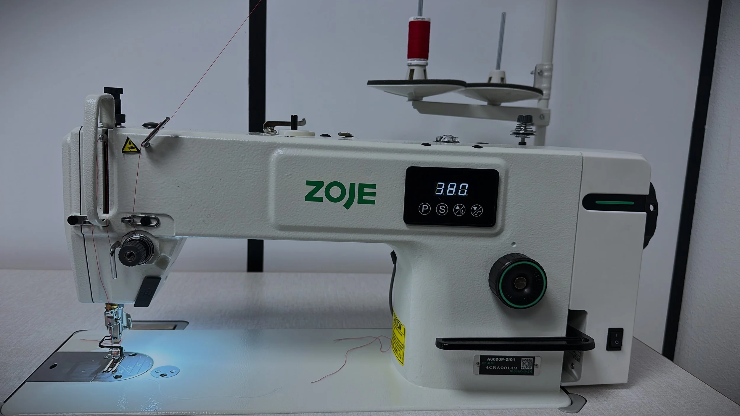 Máquina de coser blanca con pantalla digital y devanado de hilo rojo en la parte superior, en un ambiente de taller o costura.