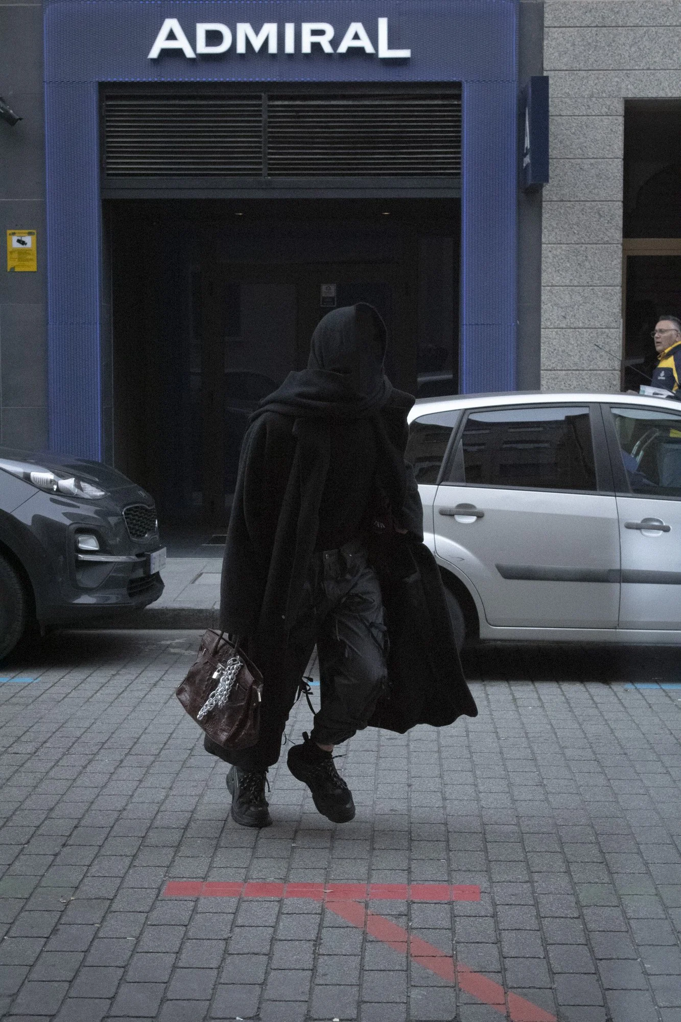 Persona con ropa negra, cargando una bolsa de mano y una mochila, cruzando en línea roja en la acera frente a un edificio azul con letrero que dice 'Admiral', y un coche gris estacionado en la calle