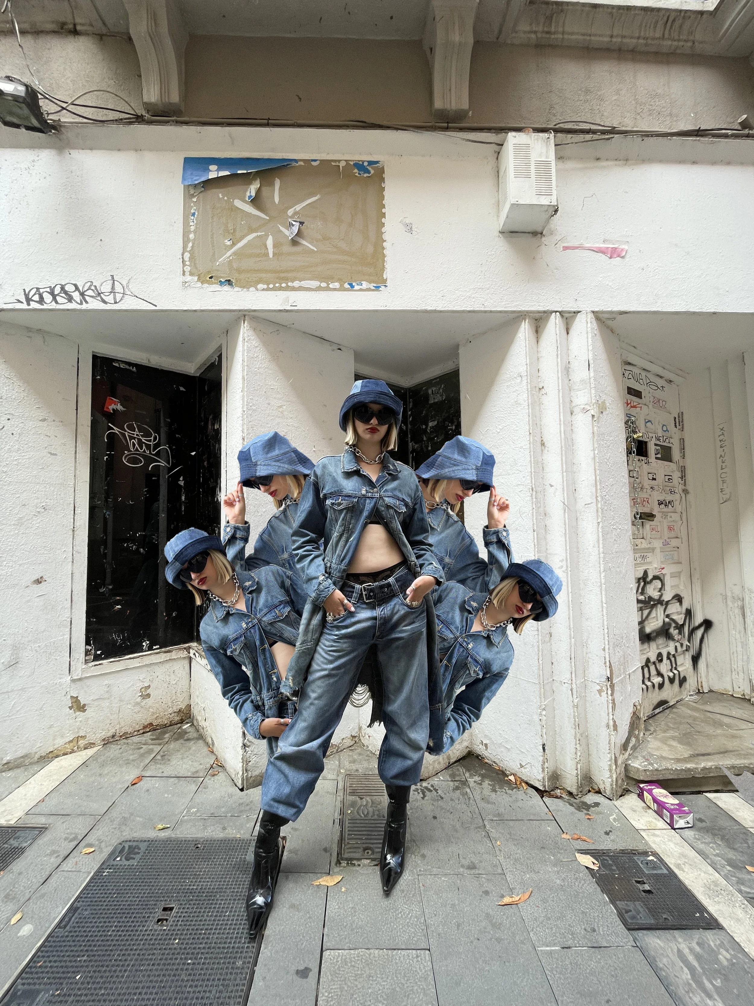 Mujer en atuendo de mezclilla con sombrero, gafas de sol y botas con tacón alto, posando en una calle urbana con graffitis y graffiti en las paredes, rodeada de varias versiones de sí misma en diferentes posiciones.