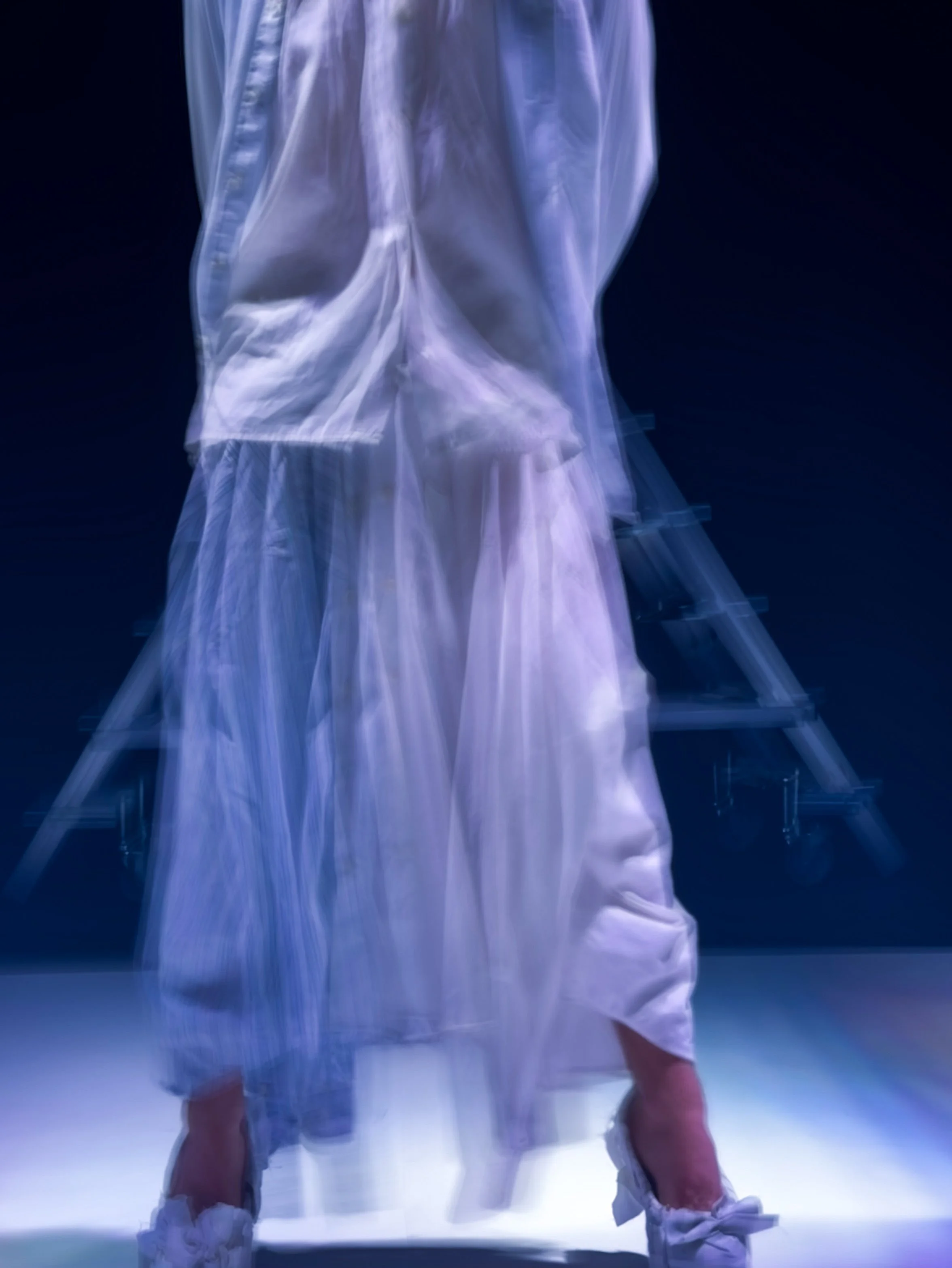 Figura humana vestida con ropa de color blanco, de espaldas, en un escenario oscuro, con iluminación que resalta la tela y las piernas.