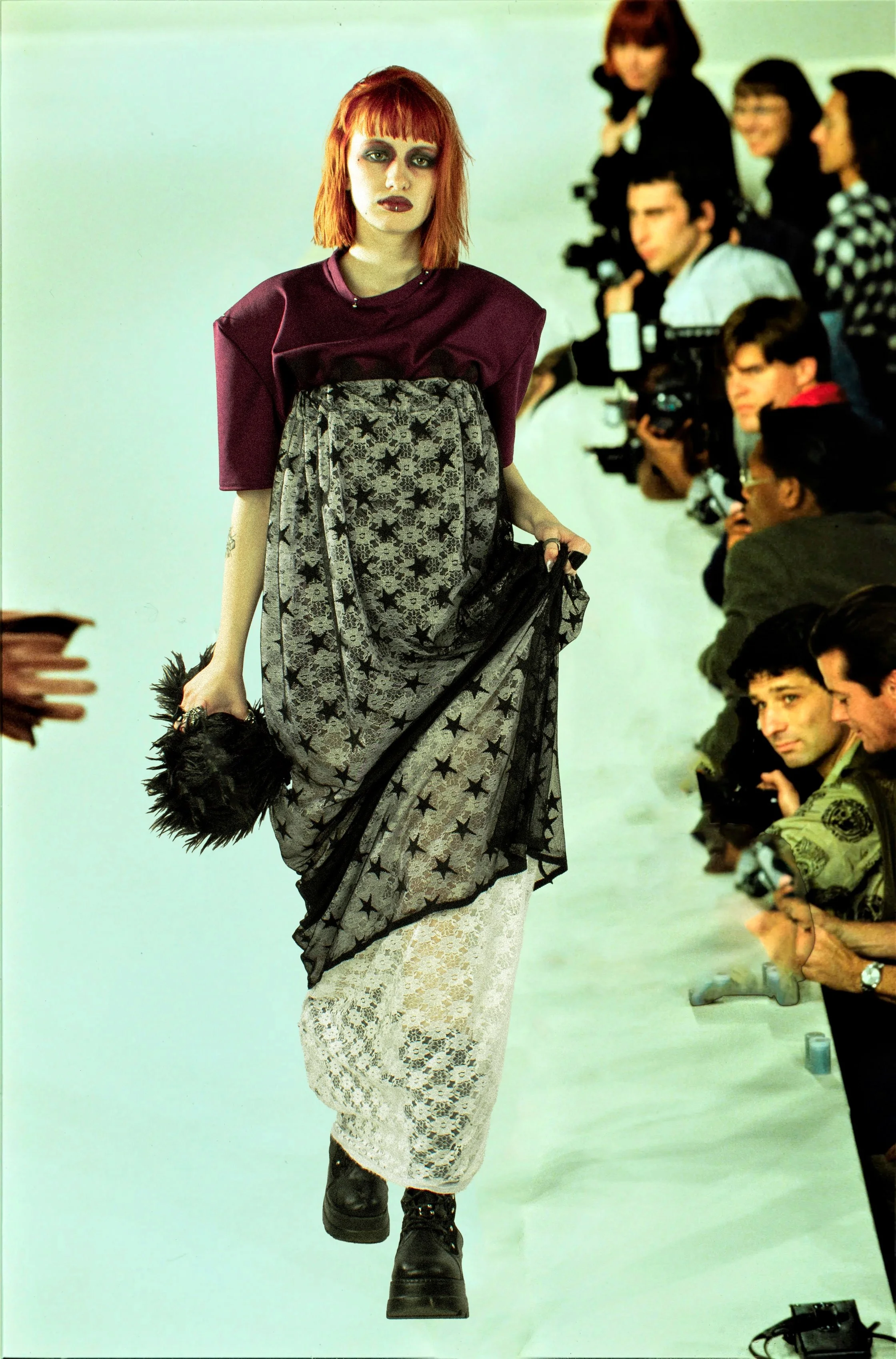 Modelo con cabello rojo y maquillaje oscuro caminando en pasarela con público a un lado, llevando un vestido largo y gabardina con falda de encaje y botas negras.