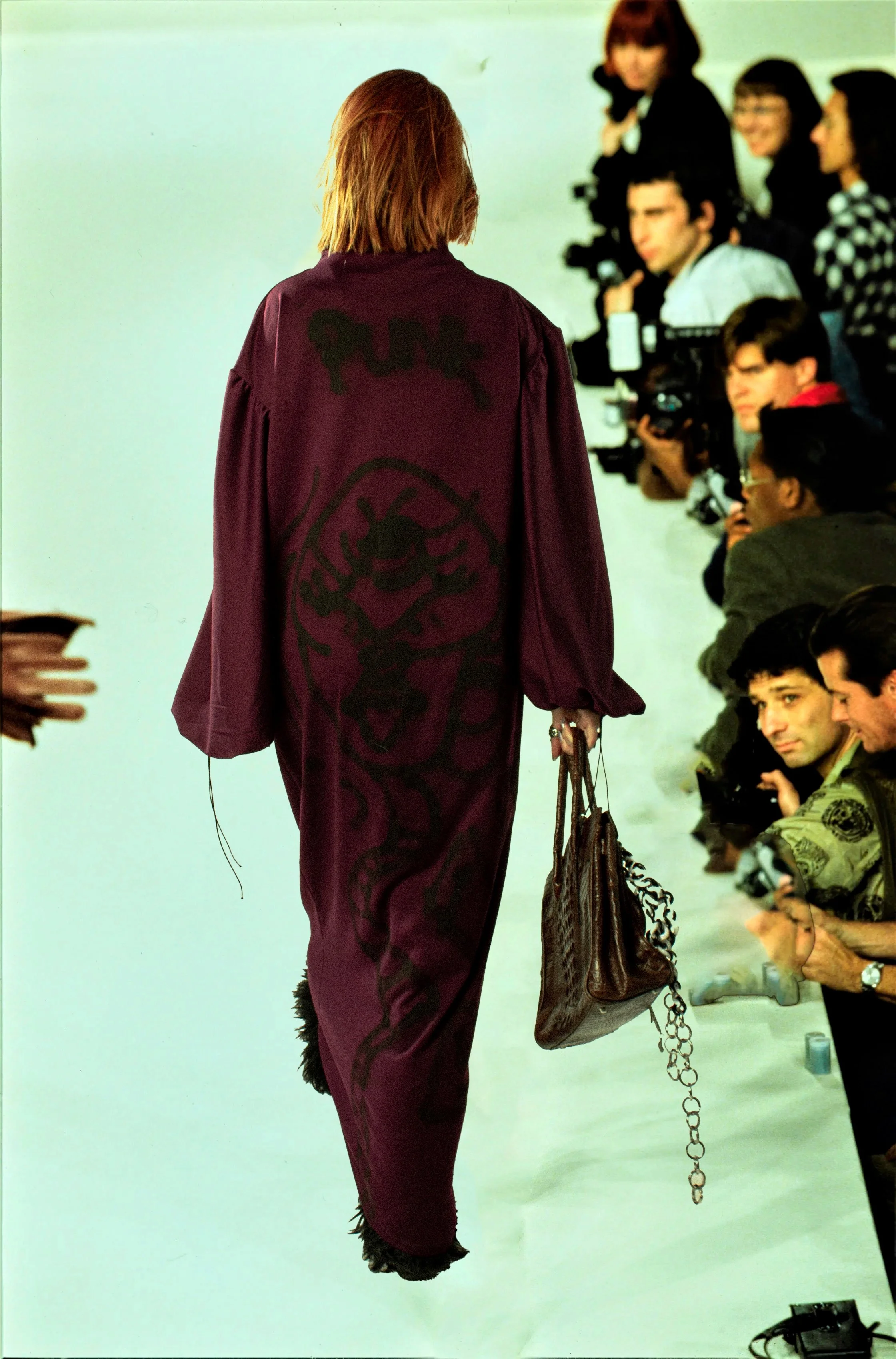 Persona con cabello rubio y largo, viste una túnica morada con un diseño oscuro en la parte trasera, y lleva un bolso con cadena, observada desde atrás, en lo que parece ser un evento de moda o una presentación con varias personas y cámaras en el fondo.