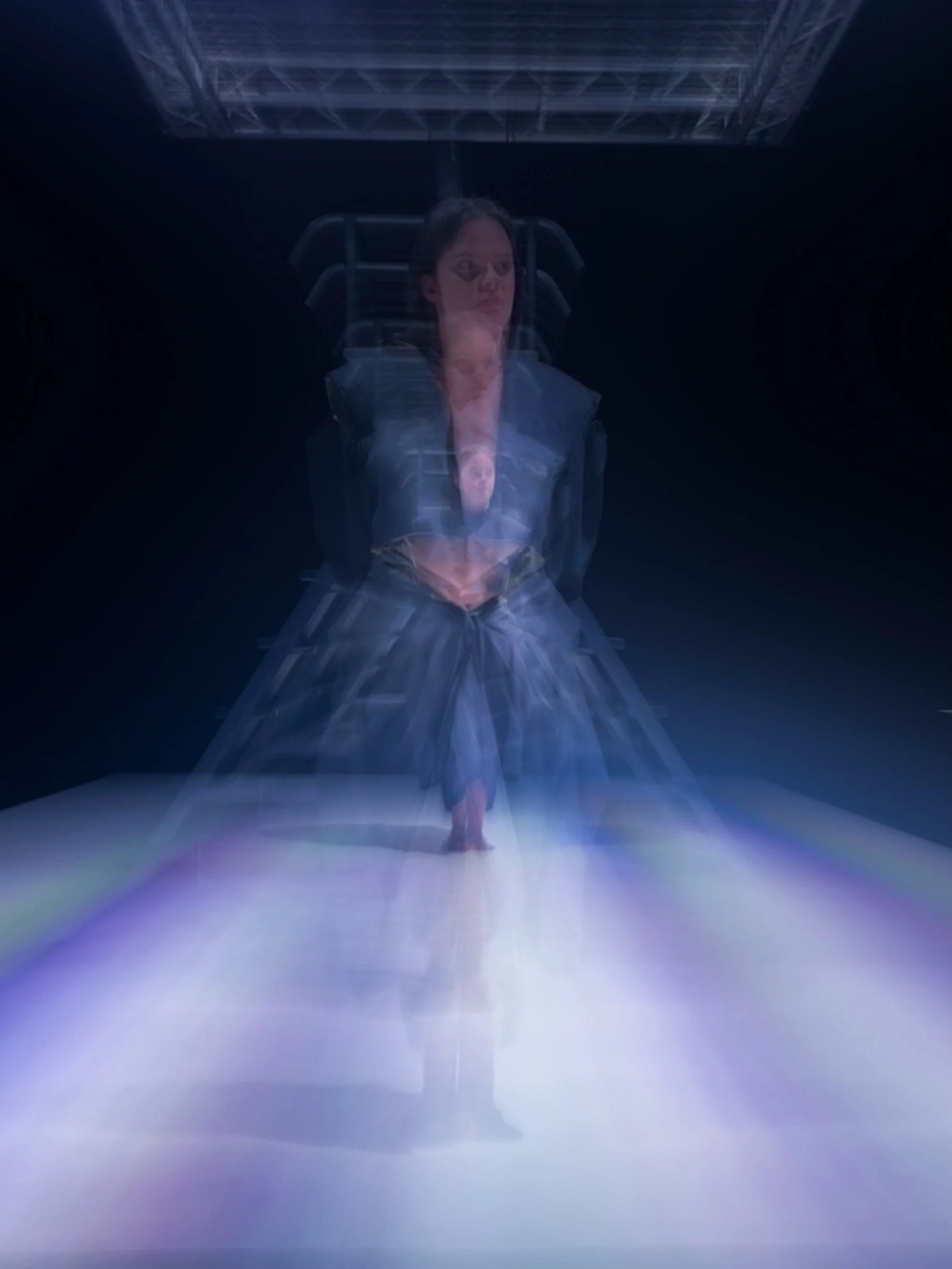 Una mujer con cabello oscuro sentado en una silla en un entorno oscuro, con efectos de movimiento y luz que crean una imagen distorsionada y surrealista.