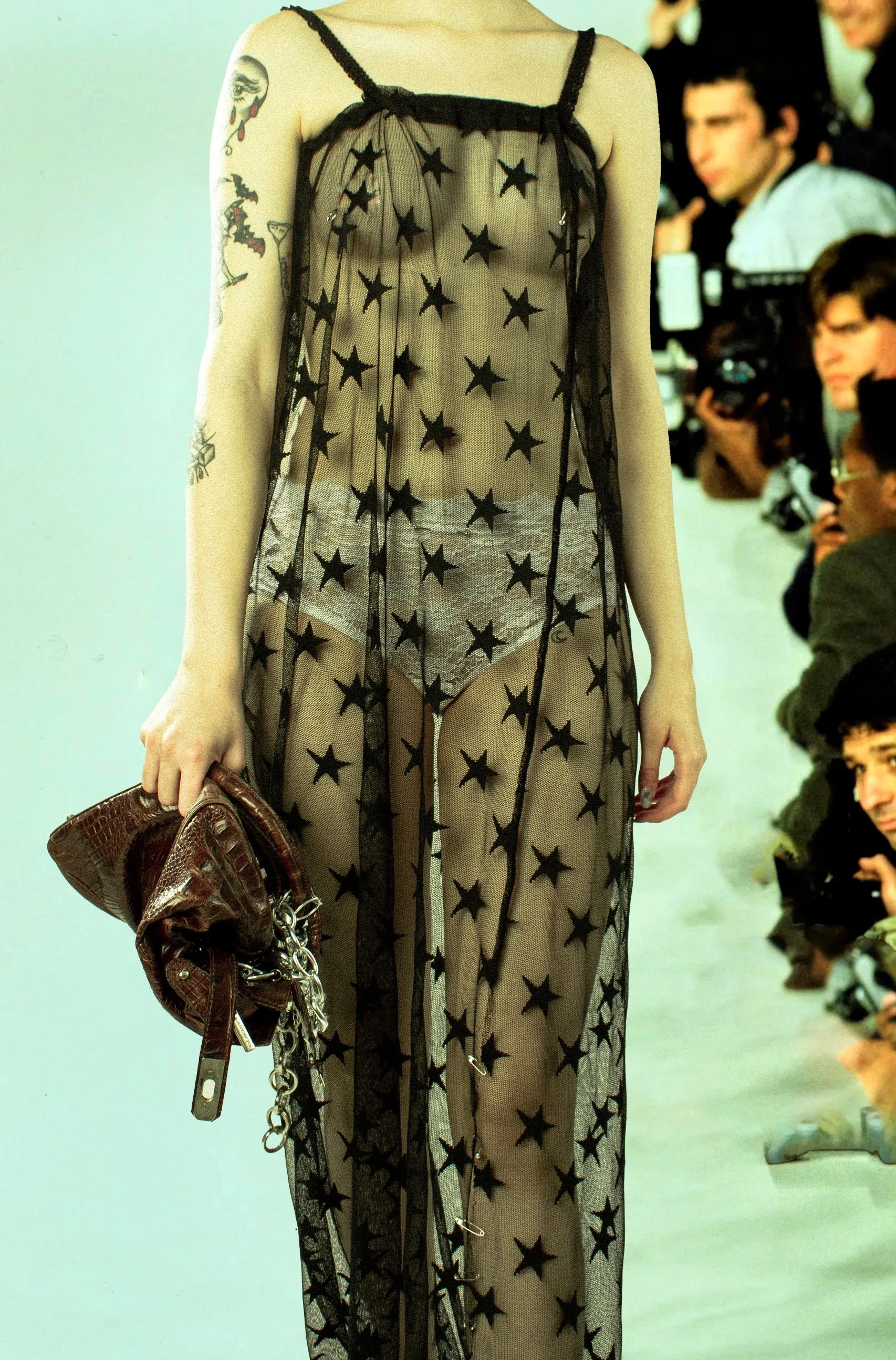 Vestido largo de malla transparente con estampado de estrellas negras, llevado por una modelo en una pasarela, con tatuajes visibles en el brazo izquierdo y sosteniendo un bolso de cuero marrón.