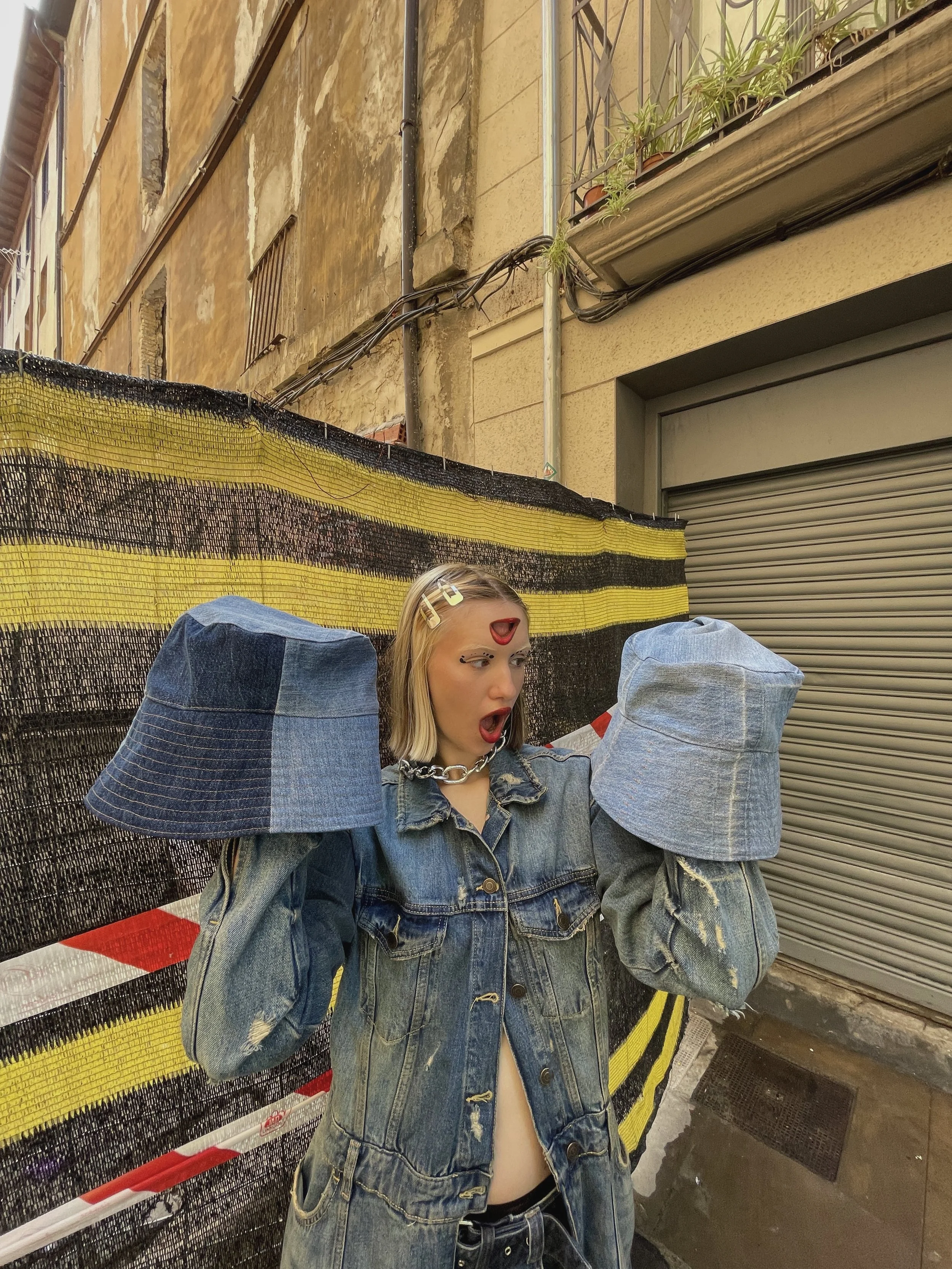 Mujer con cabello rubio, gafas de sol, chaqueta de mezclilla y cinturilla de pantalones, con boca abierta y expresión de sorpresa, en un entorno urbano con paredes envejecidas y una cortina de seguridad.