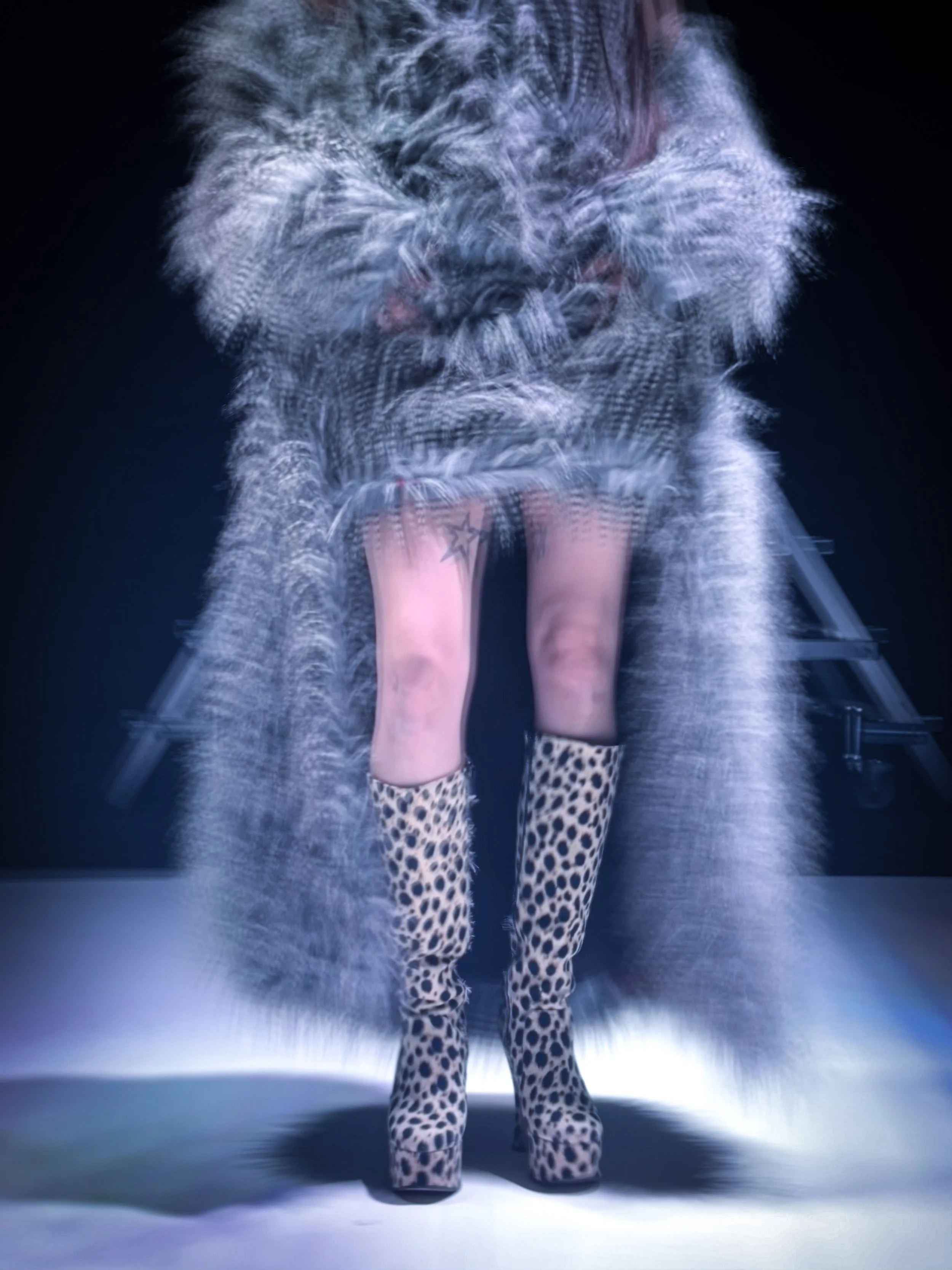 Persona con pantalones de leopardo y botas negras con plataforma elevada, de espaldas, en un escenario oscuro con iluminación modernista.
