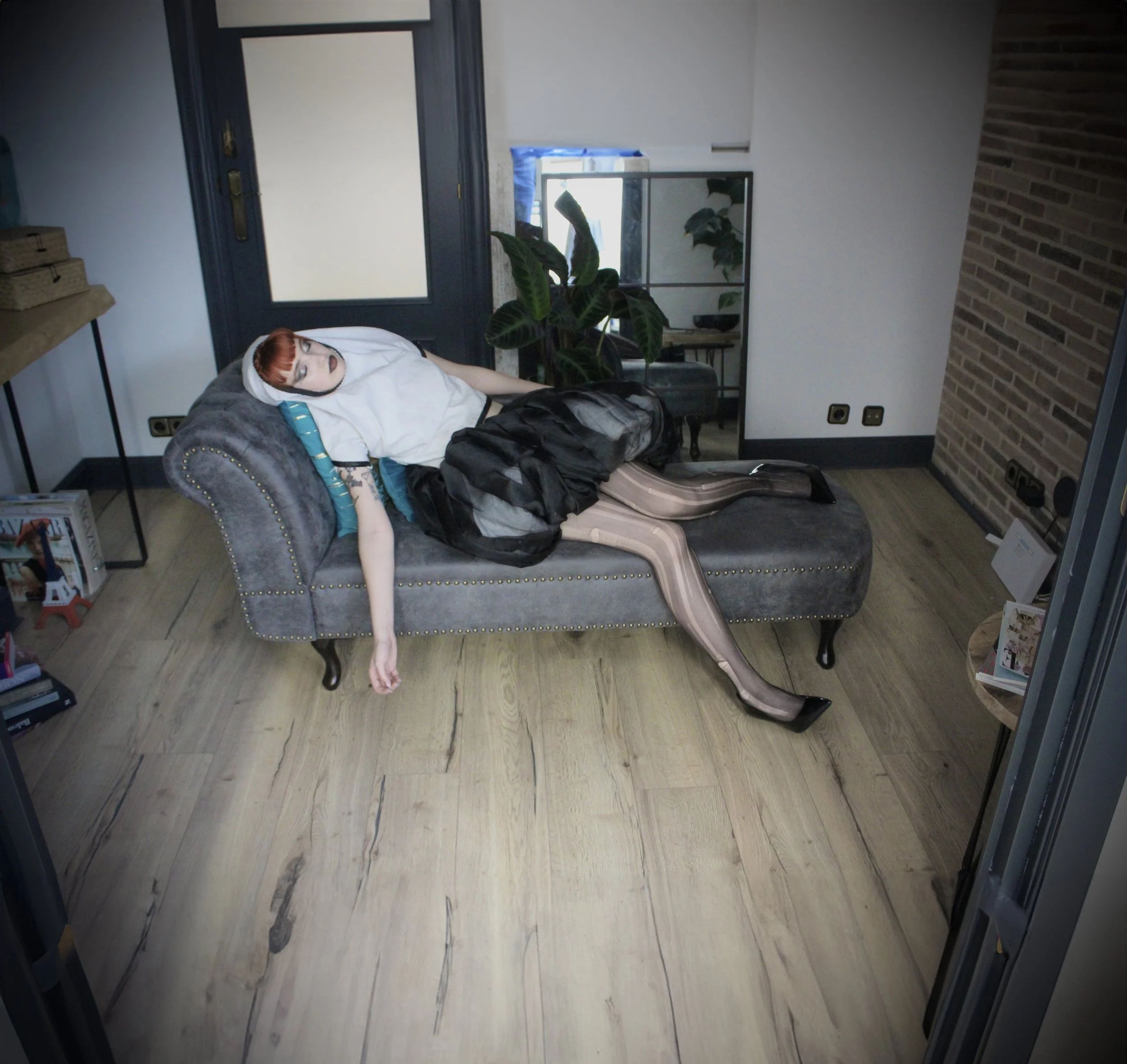 Interior de una habitación con un sillón gris, plantas verdes, una mesa de madera, un espejo, una ventana con cortinas azules y paredes blancas. Incluye algunos libros y juguetes en la esquina.