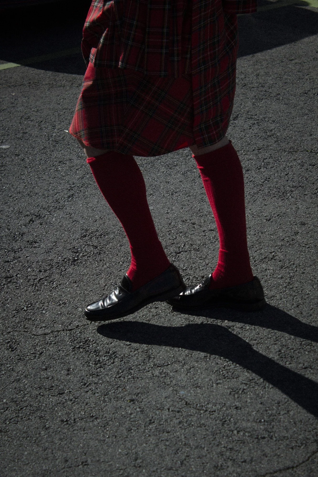 Piernas de una persona con falda escocesa roja y negra, medias rojas y zapatos negros en asfalto con sombra