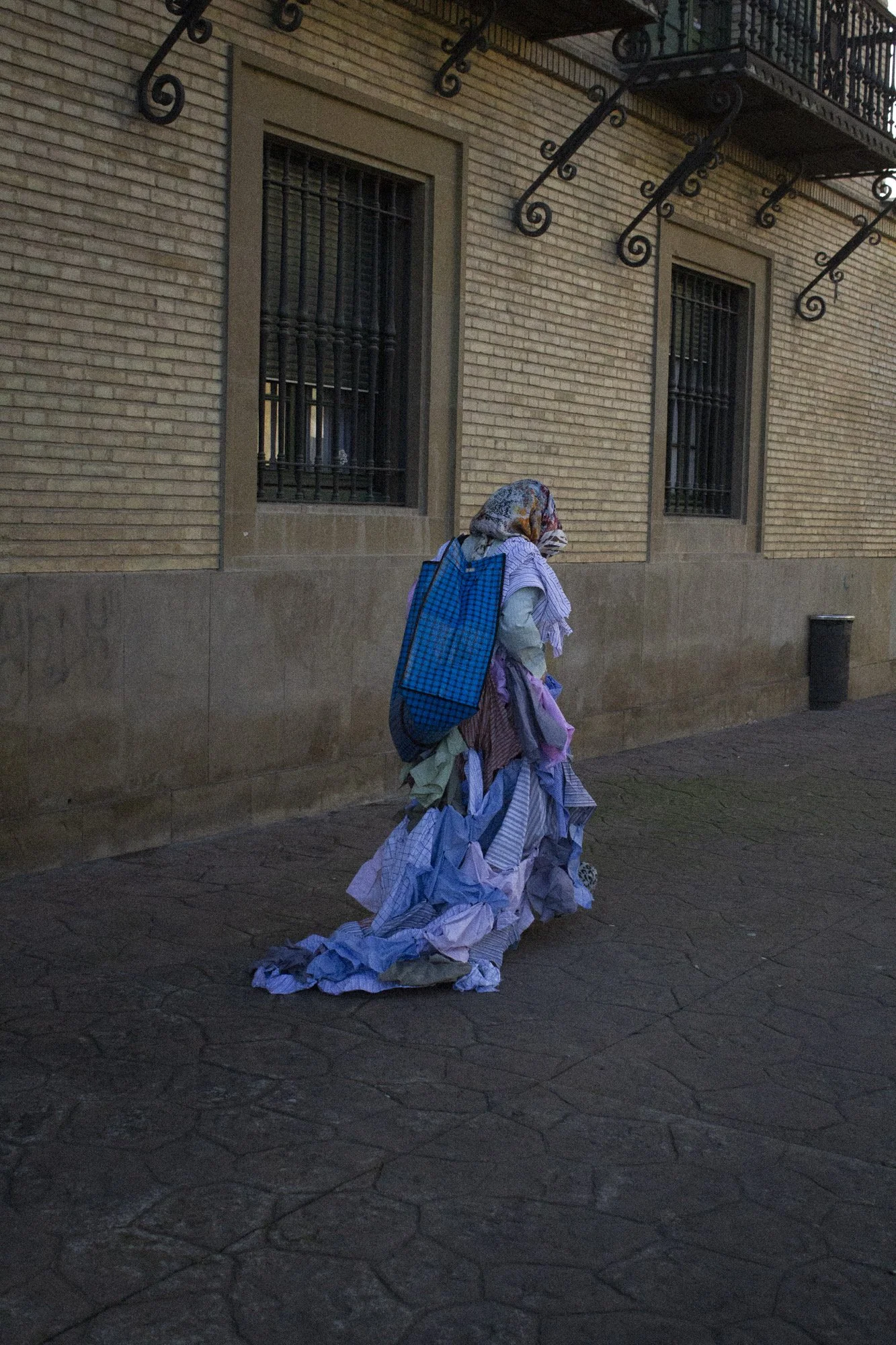 Mujer vestida con ropa de colores claros y una bufanda, llevando un saco grande de tela azul lleno de papeles o prendas, caminando por la acera de una calle urbana con un muro de ladrillos y ventanas con rejas