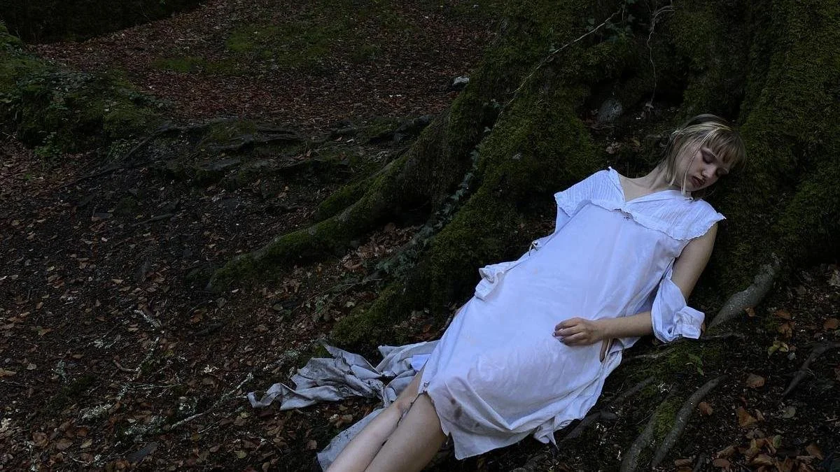 Mujer con vestido blanco acostada en el suelo de un bosque, parece estar soñando o durmiendo.