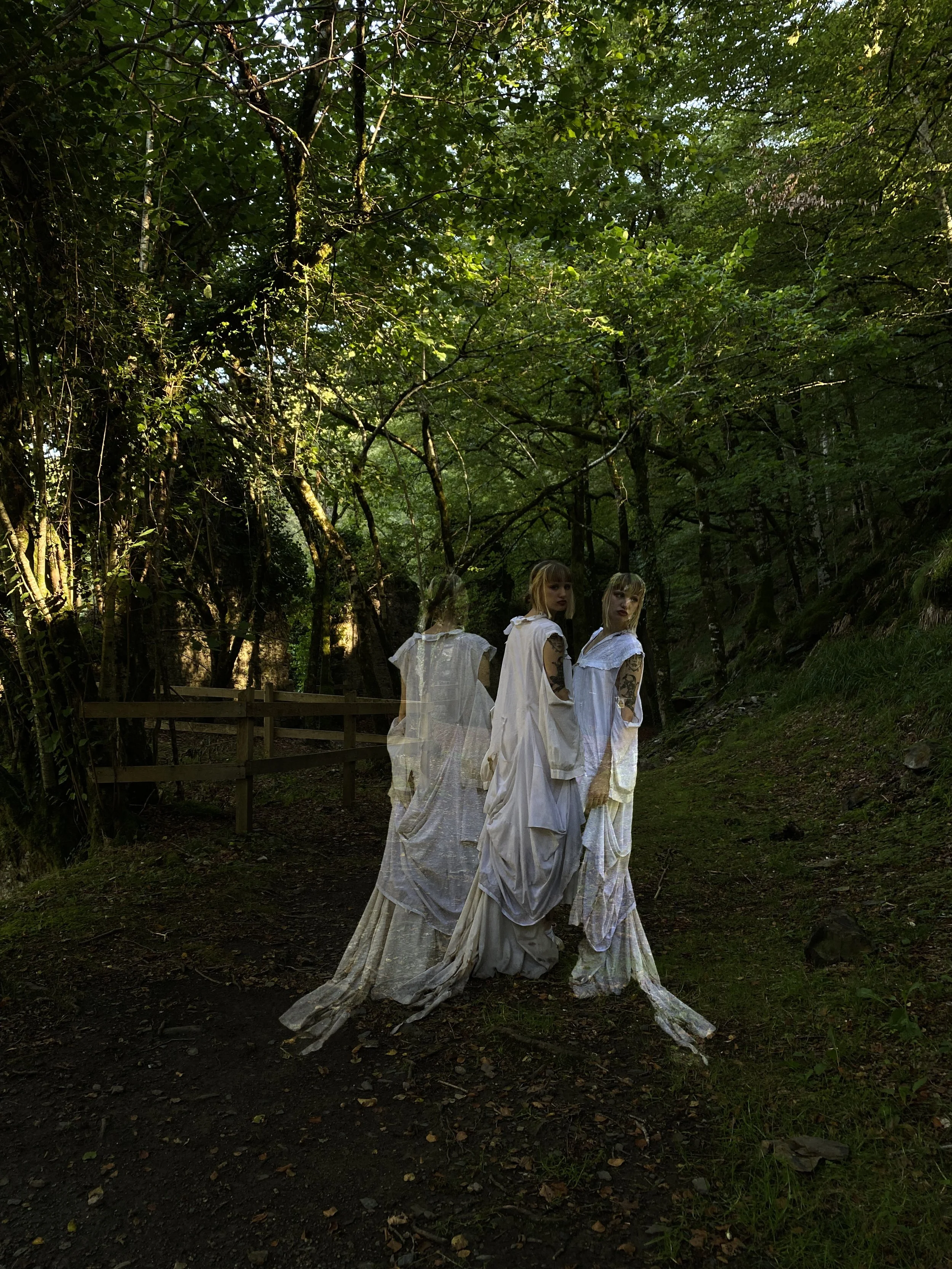 Tres mujeres vestidas con ropa blanca y de aspecto elegante se encuentran en un entorno boscoso, con árboles verdes y un sendero de tierra.