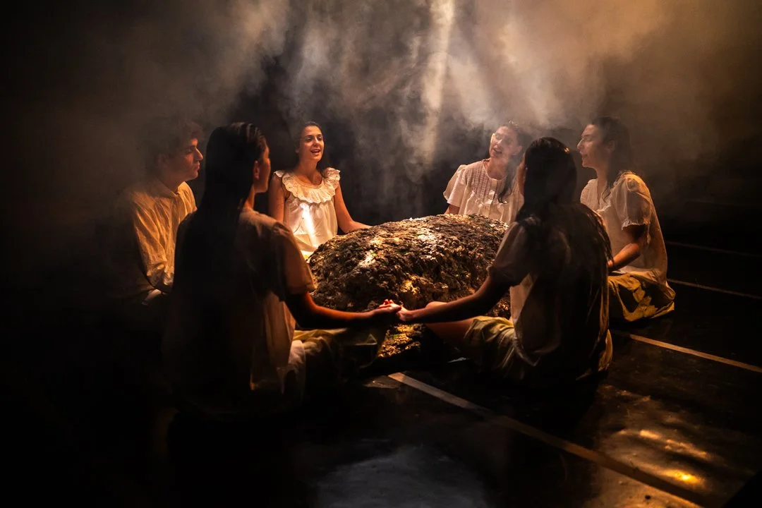 Grupo de siete personas sentadas en círculo alrededor de una roca en un lugar oscuro y con humo o vapor, algunos parecen estar riendo o conversando.