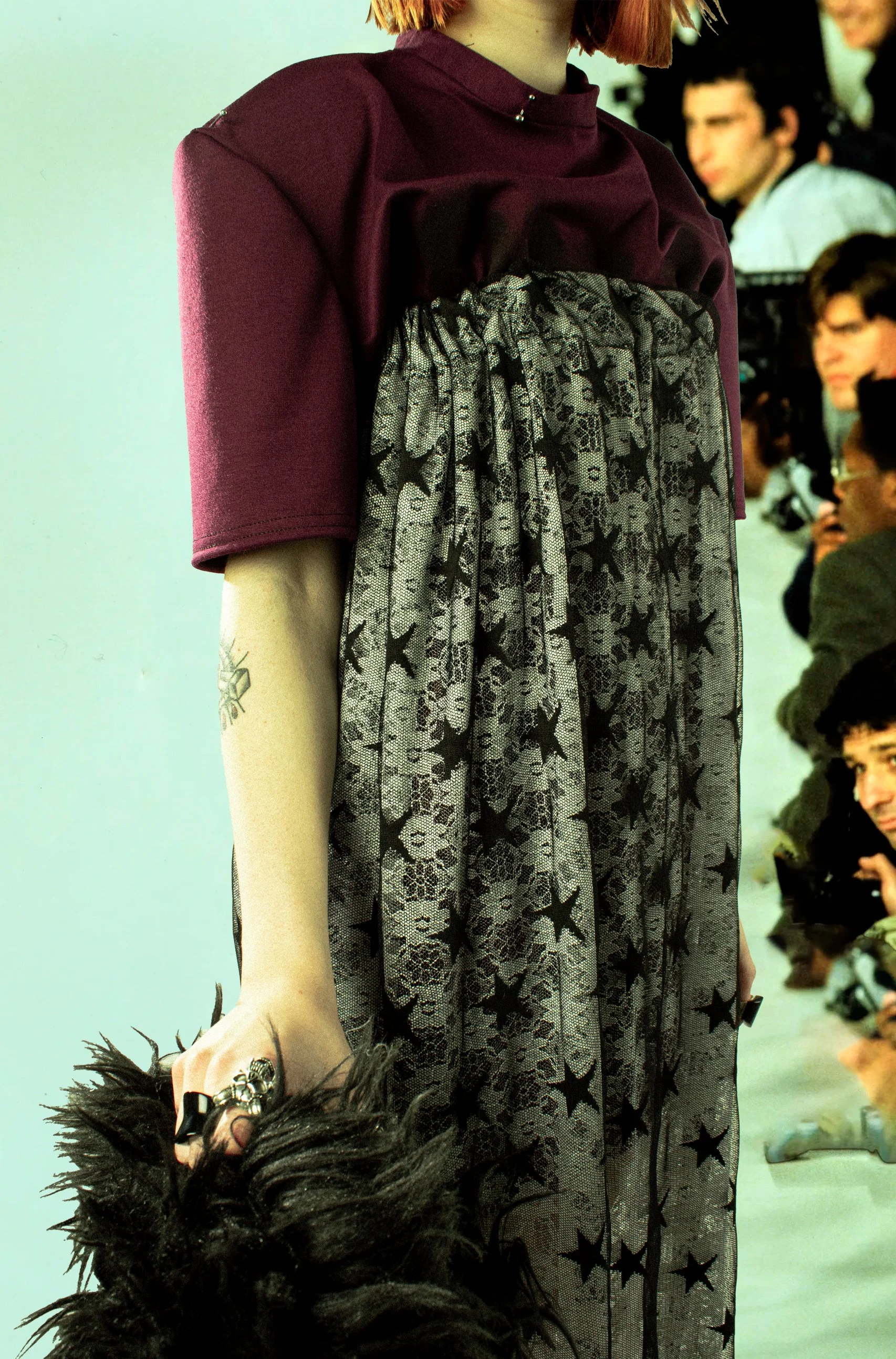 Mujer con blusa morada, falda de encaje con estrellas negras, lleva pena de mano con plumas y accesorios en la mano, en un entorno de pasarela o moda.