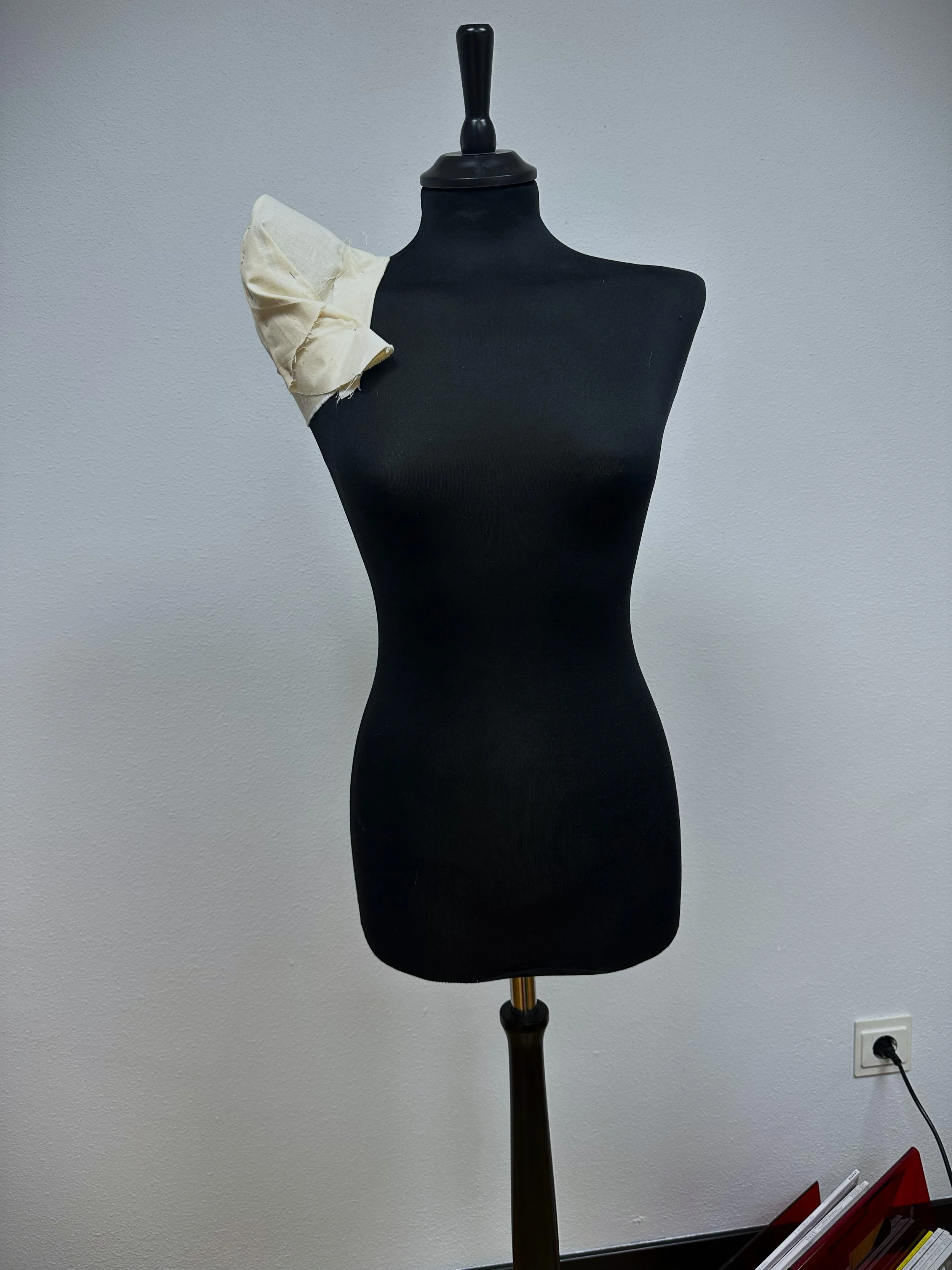 Maniquí negro sin cabeza con soporte, con un trozo de tela blanca en el hombro izquierdo, en una pared blanca y un enchufe en la parte inferior derecha.
