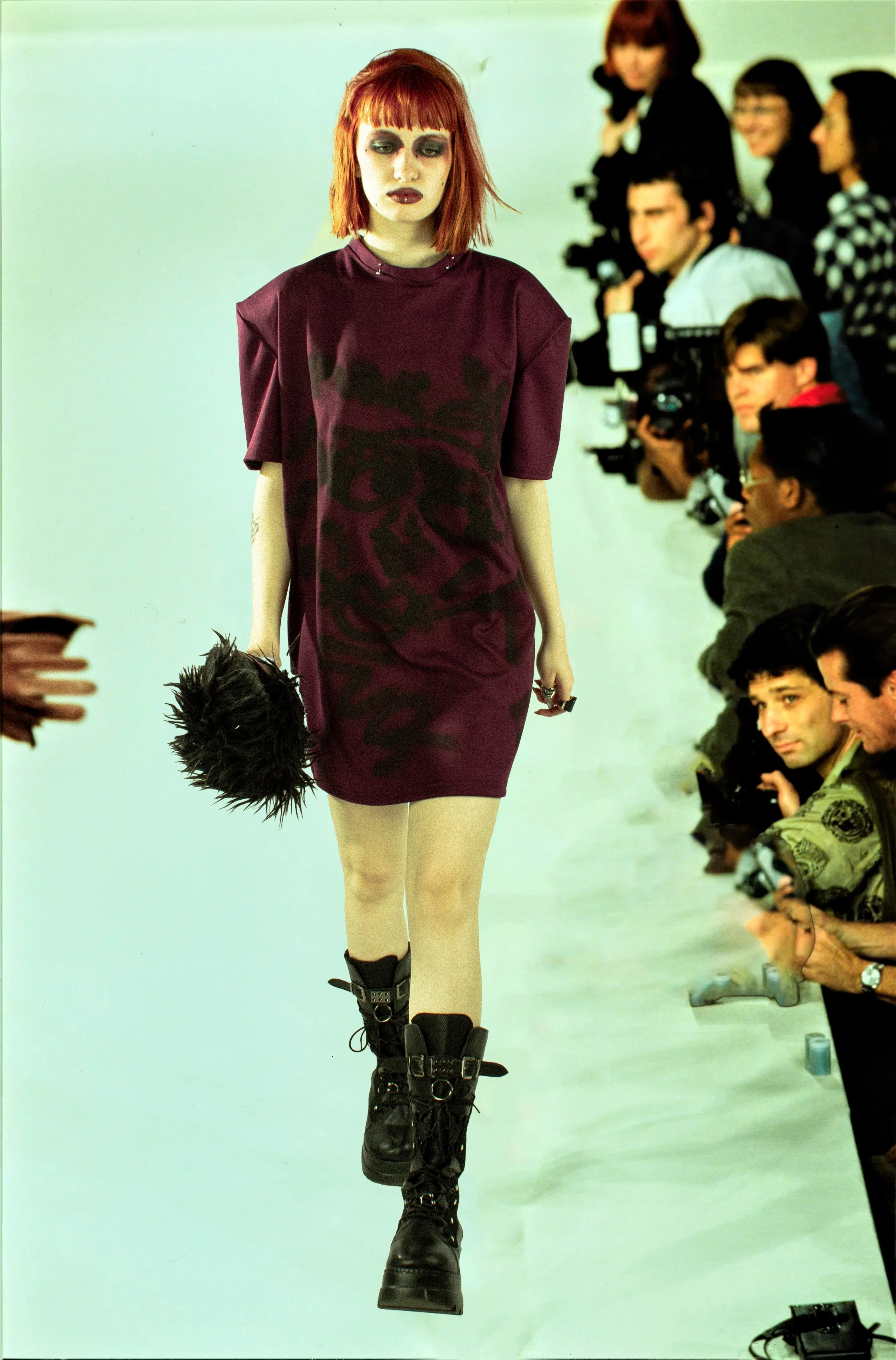 Mujer con cabello rojo, vestida con ropa oscura y botas robustas, caminando en una pasarela frente a fotógrafos y espectadores.
