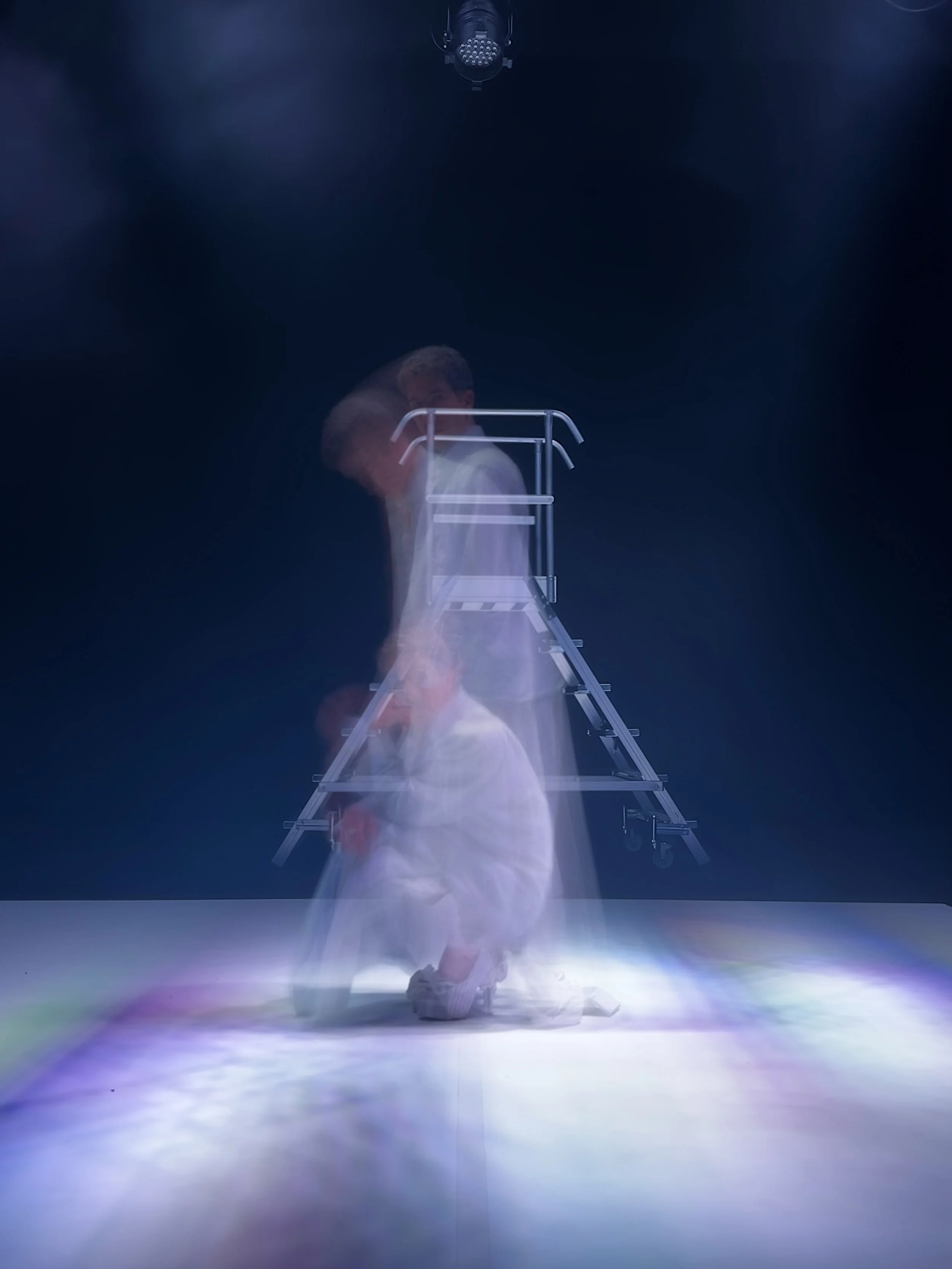 Persona en ropa blanca en posición de yoga, apoyada en las rodillas y con las manos en el suelo, con una escalera metálica delante y un fondo negro, con iluminación centrada.