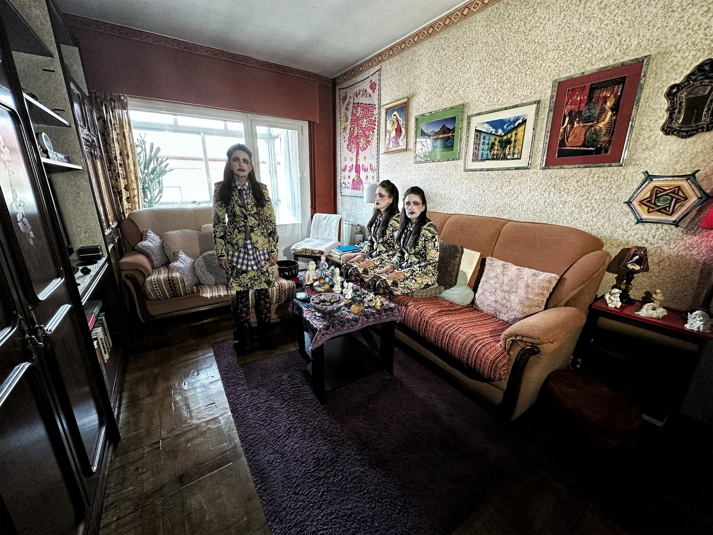 Cuatro personas con vestuario de estilo tradicional y maquillaje oscuro en un salón decorado con cuadros y tapices, sentadas y una más de pie.