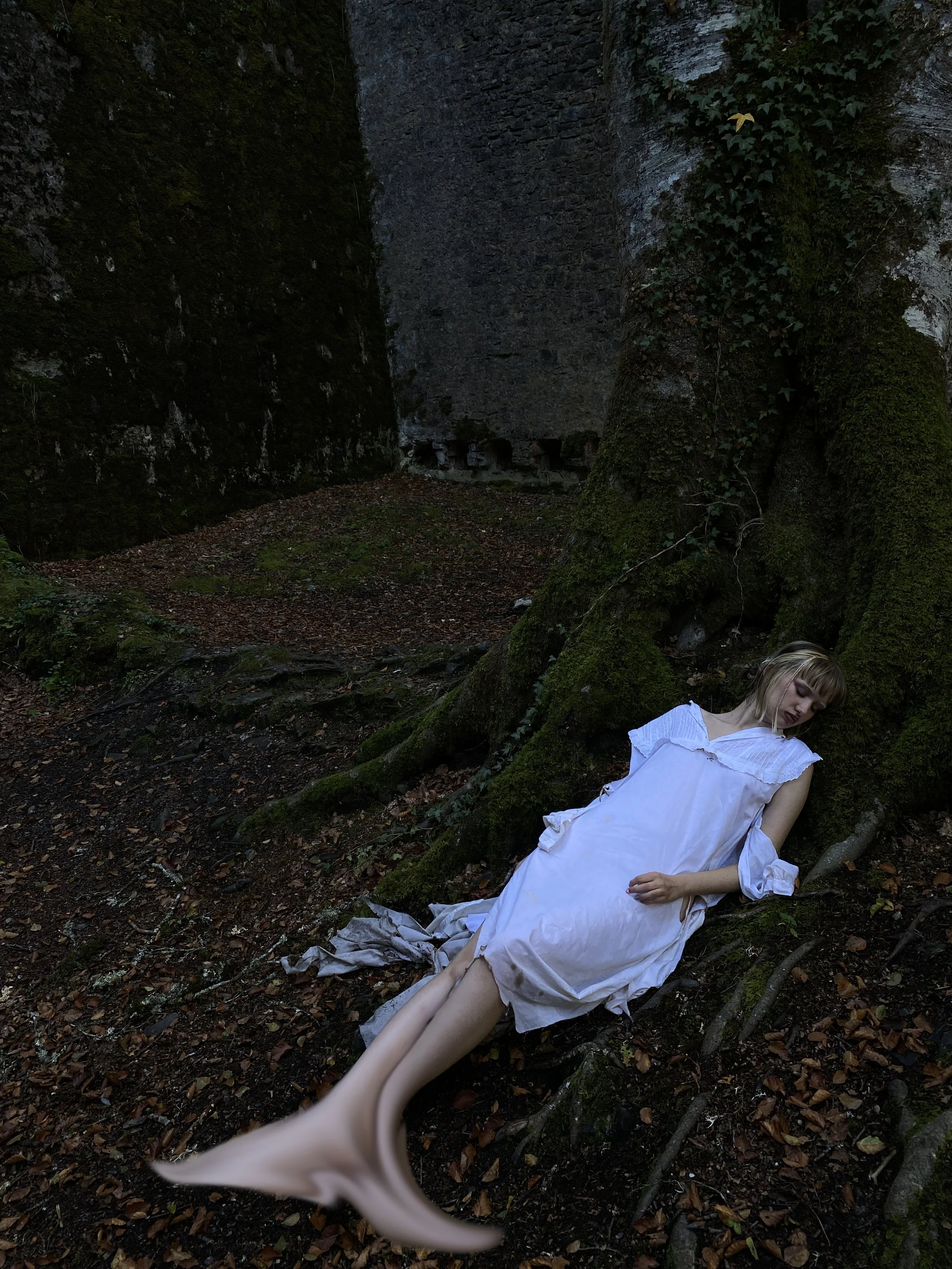 Mujer acostada sobre raíces y hojas en un bosque, vestida con un vestido blanco, con una expresión pensativa o tranquila.