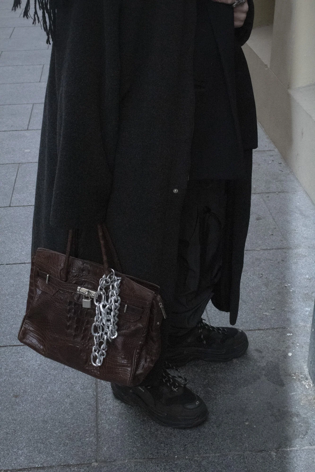 Persona con ropa negra que lleva un bolso de mano marrón con cadenas plateadas, zapatos negros, y pantalones oscuros, de pie sobre un pavimento de piedra.