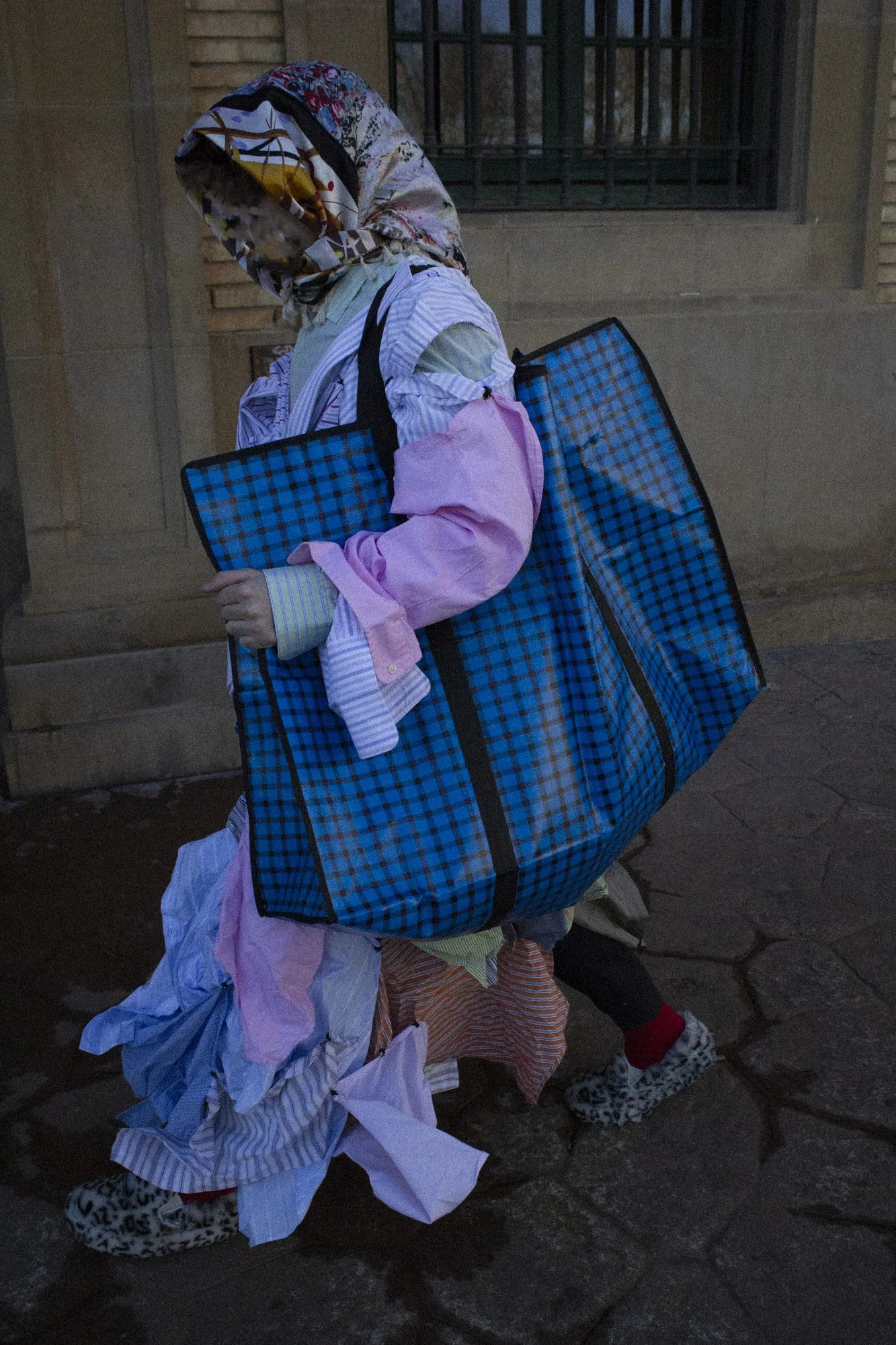 Persona con ropa multicolores y una bufanda con capucha, cargando una segunda bolsa gigante con un andador, caminando en la calle frente a un edificio con rejas en la ventana.
