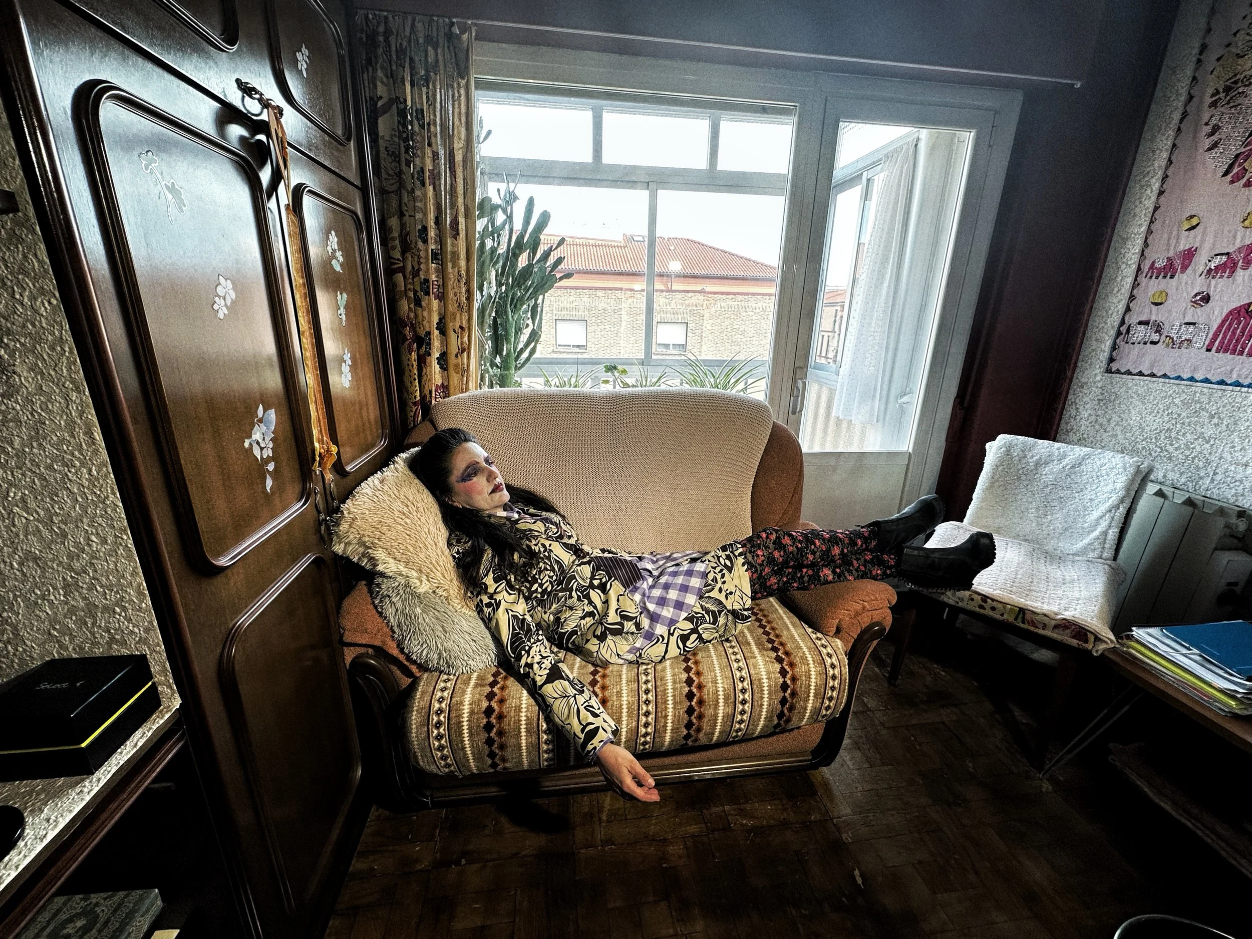 Una mujer con maquillaje llamativo y ropa de colores oscuros, acostada en un sillón en un ambiente interior con ventana y plantas.